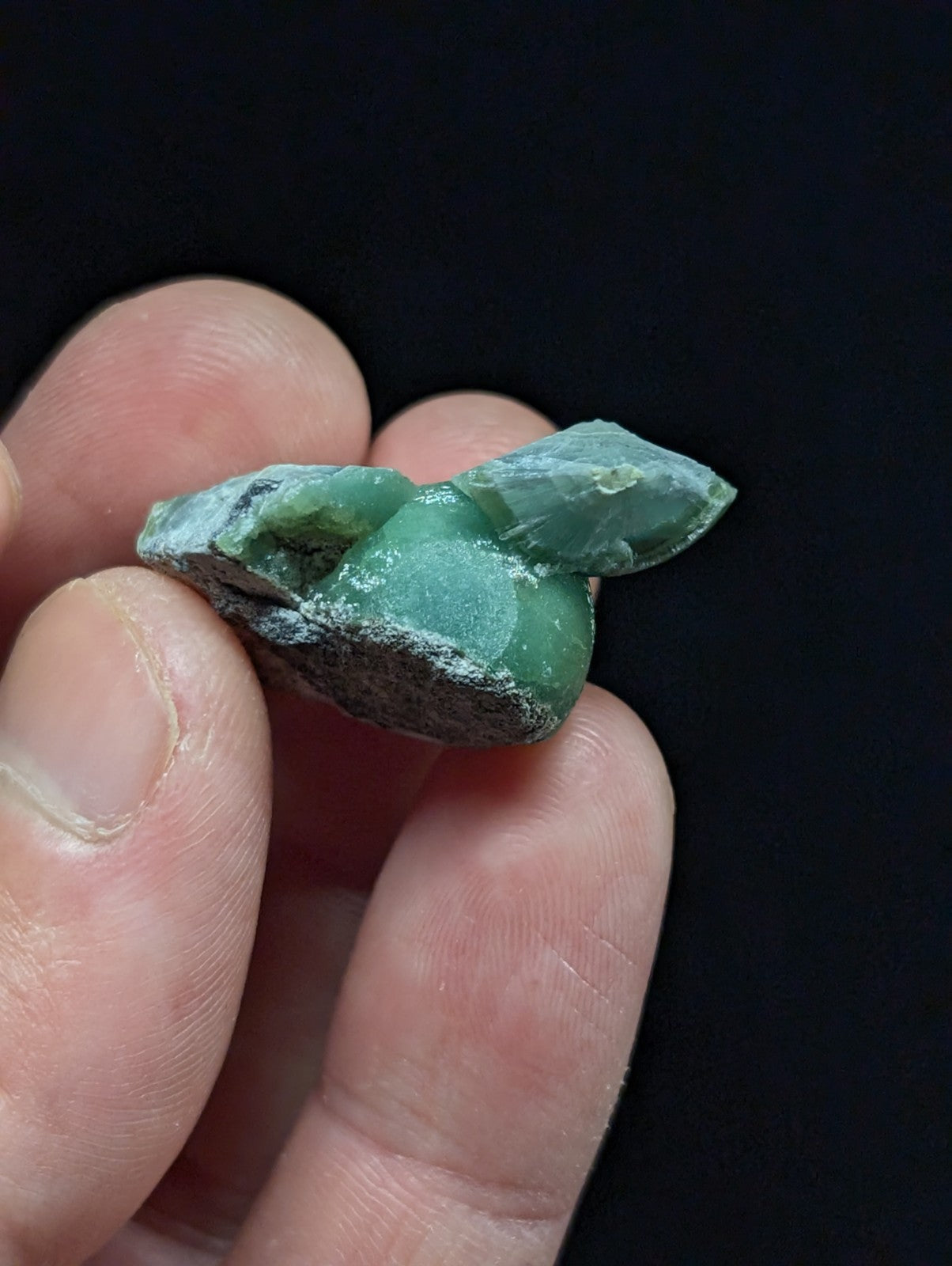 A hand holds a small, irregularly shaped green jadeite mineral specimen with visible crystalline formations and some dark matrix material.