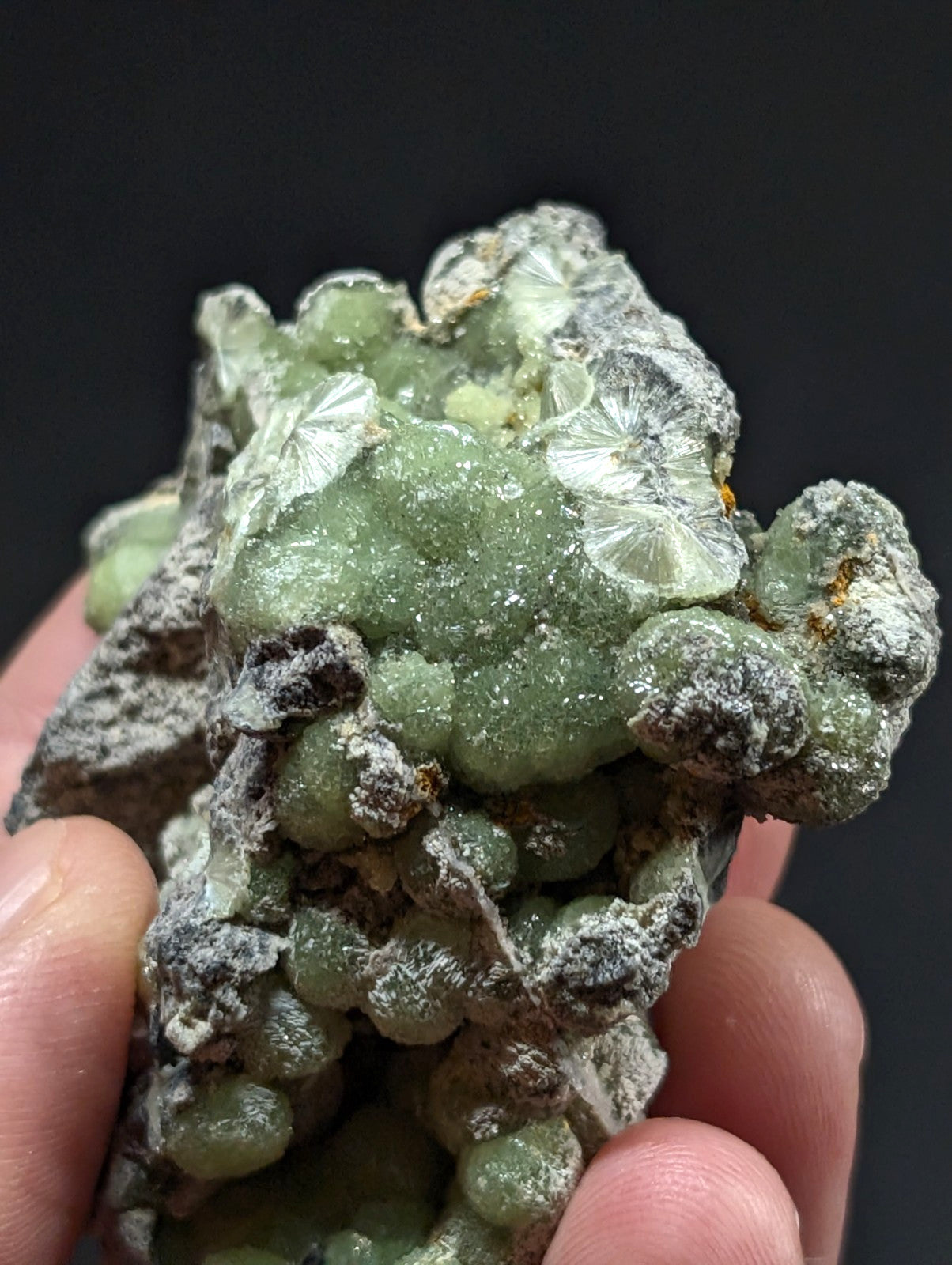 A hand holds a green, translucent mineral specimen with crystalline formations and grayish matrix.