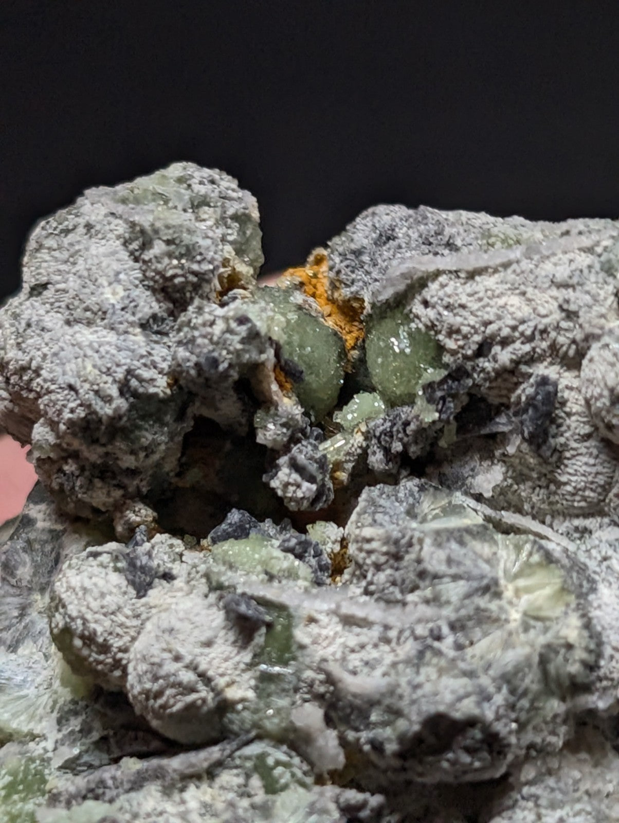 A rough, grayish-white mineral specimen with embedded translucent green crystals and patches of orange-yellow material.