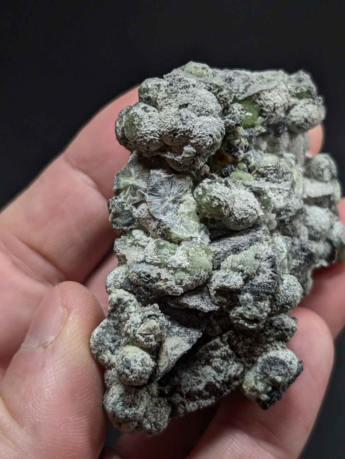 A hand holds a rough, clustered mineral specimen with greenish translucent crystals embedded in a gray, powdery matrix.