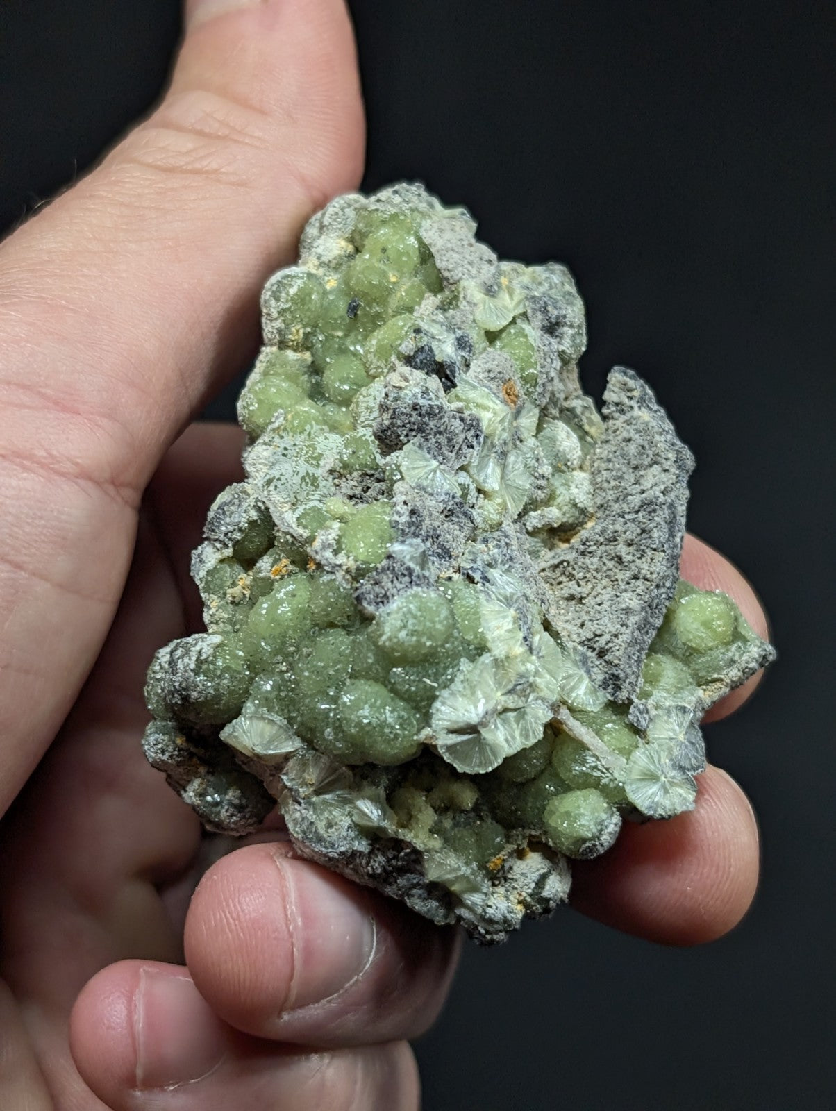 A hand holds a cluster of green, spherical mineral formations with translucent, leaf-like crystals embedded in a grayish matrix.