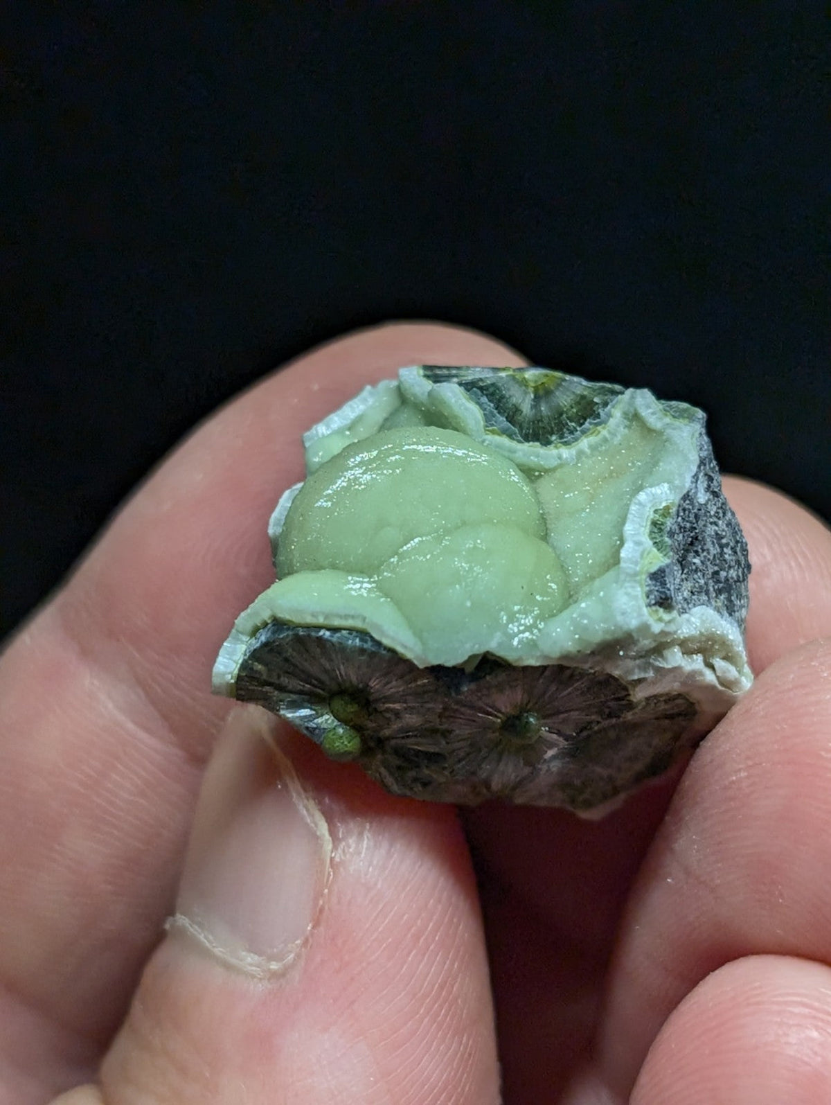 A hand holds a natural, irregularly shaped mineral specimen featuring a vibrant green, glossy interior with a rougher, darker grayish-black exterior.