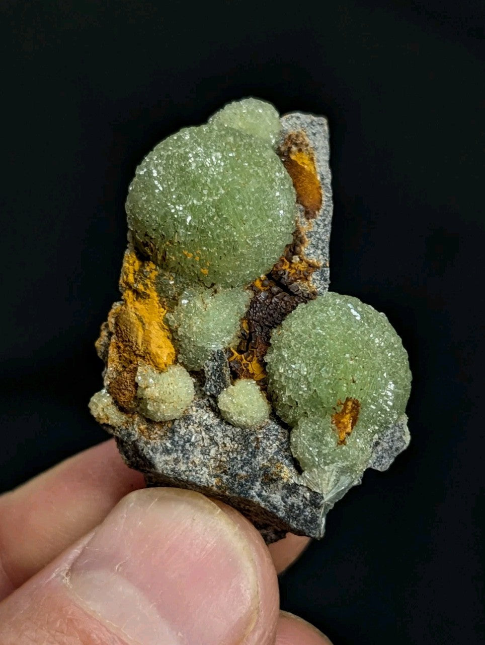 A cluster of translucent green crystals with a crystalline texture, accented by patches of orange and brown mineralization on a dark gray rock base.