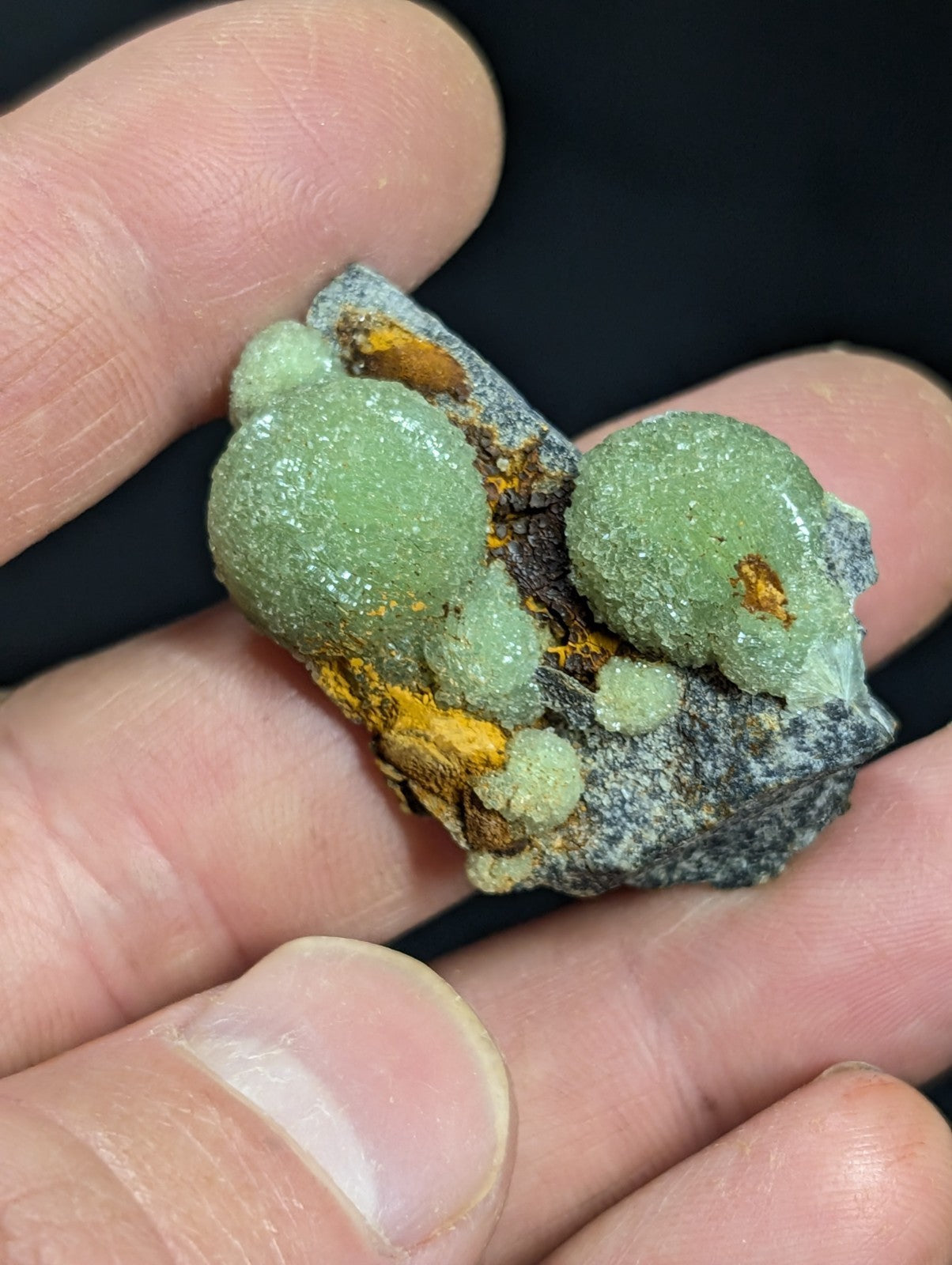 A hand holds a small, natural mineral specimen featuring vibrant green, rounded crystals growing from a dark gray rock matrix with patches of orange-yellow mineralization.