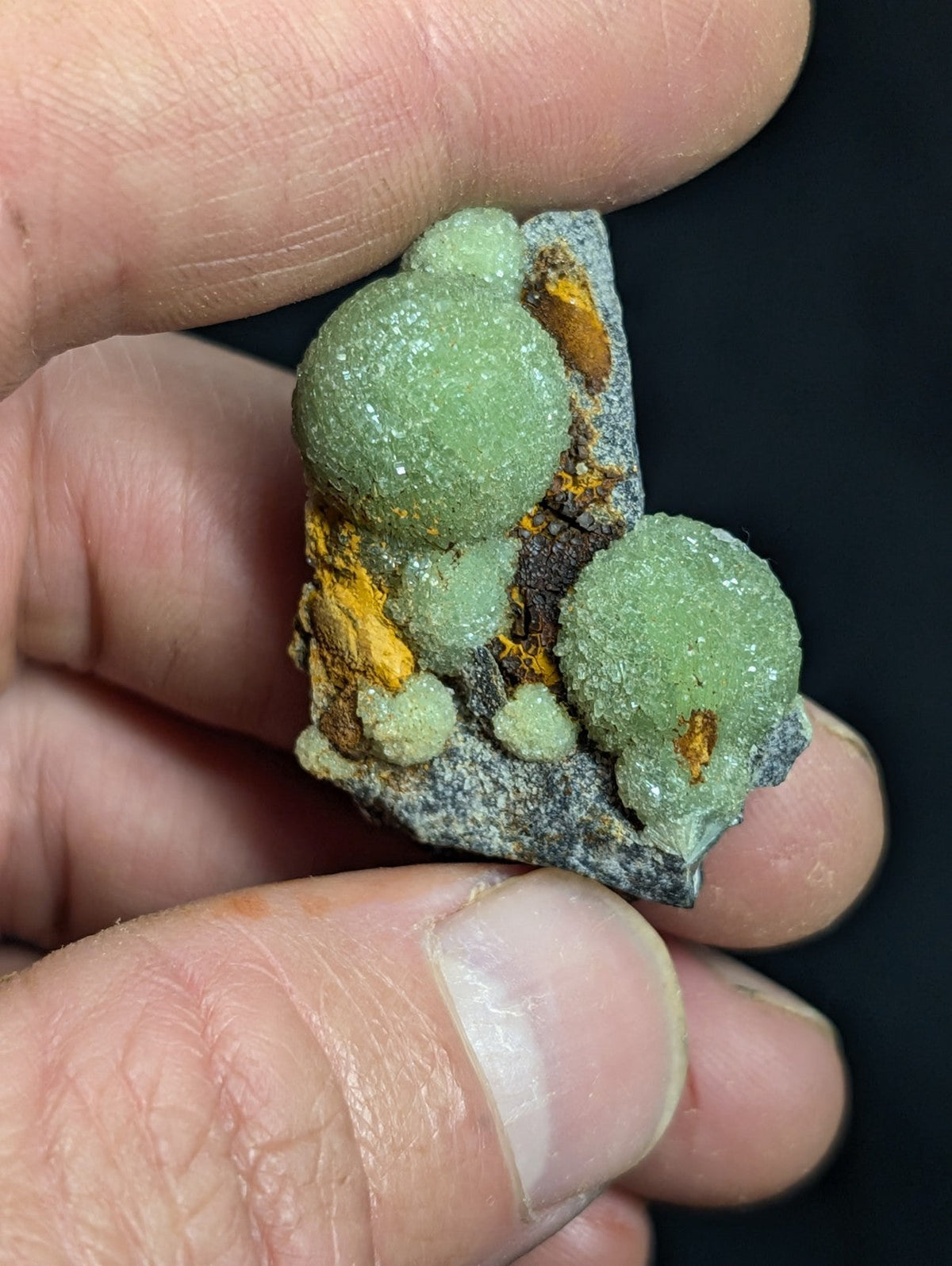 A hand holds a mineral specimen featuring vibrant green, spherical crystals with a crystalline texture growing from a dark gray rock matrix accented by patches of orange-yellow material.