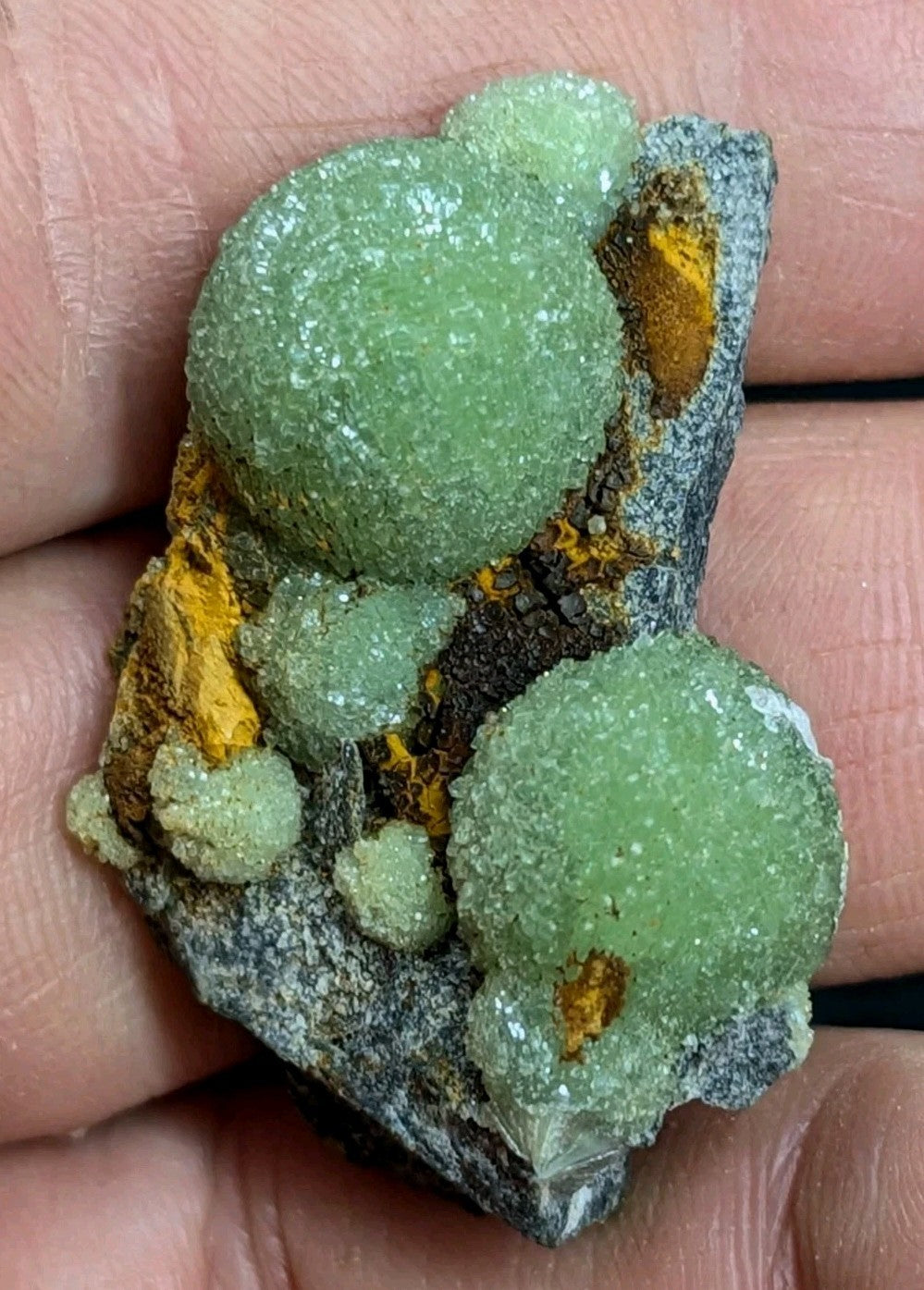 A hand holds a geological specimen featuring vibrant green, crystalline formations with a rough, textured surface and patches of orange mineral inclusions on a dark gray rock base.