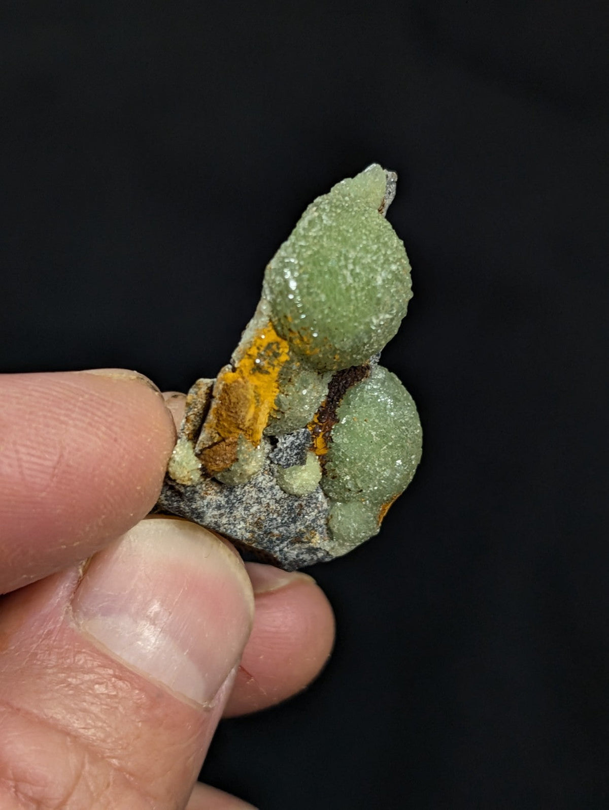 A hand holds a mineral specimen featuring two prominent green, rounded crystals with a textured surface, accented by patches of orange and gray matrix material.
