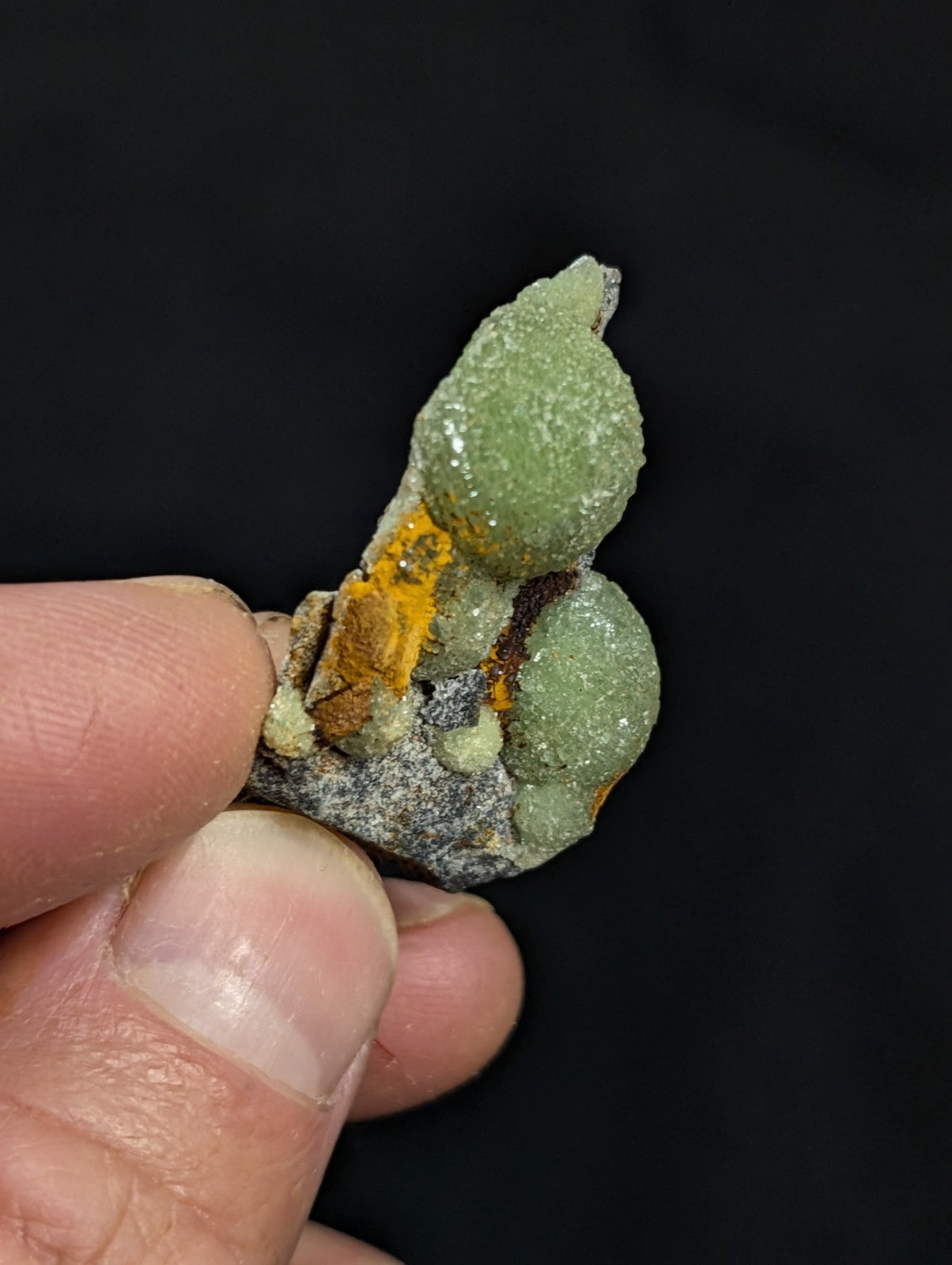 A hand holds a mineral specimen featuring two prominent green, rounded crystals with a textured surface, accented by patches of orange and gray matrix material.