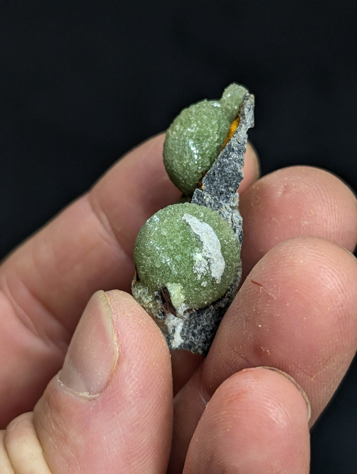 A pair of green, round, mineral-like objects with a rough texture and white patches, held between fingers.