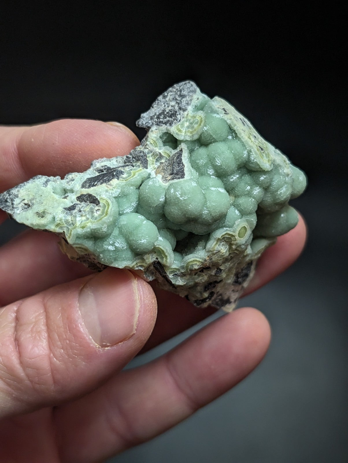 A hand holds a light green, bumpy mineral specimen with rounded nodules and darker patches.