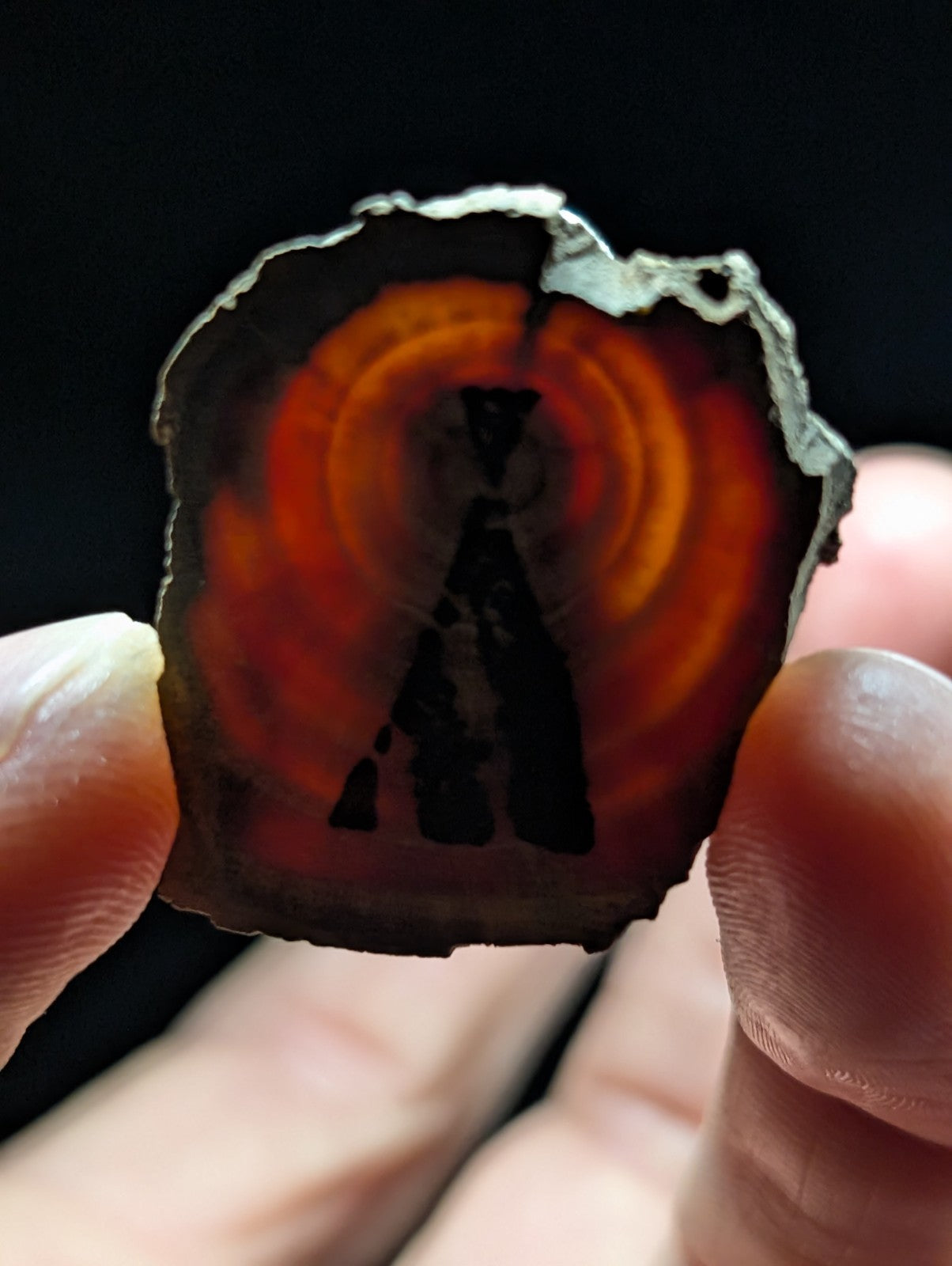 A hand holds a slice of agate with concentric orange and red bands, featuring a dark silhouette design in the center.
