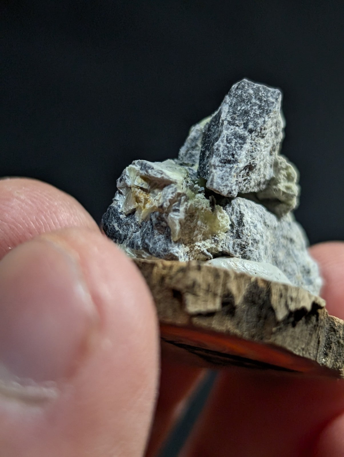 A rough, grayish mineral specimen with a speckled texture and small yellow crystalline inclusions.