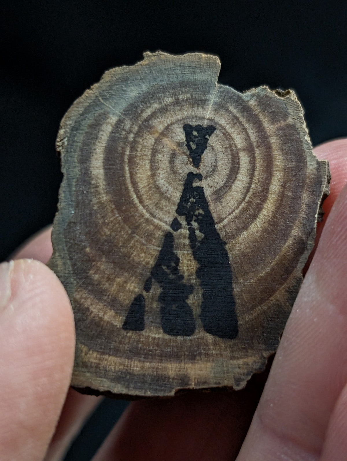 A rustic, circular wood slice with natural grain and concentric rings, featuring a dark charcoal-black silhouette of an abstract figure or symbol burned into its surface.