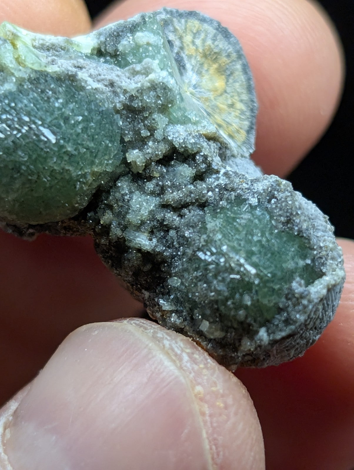 A green, crystalline mineral specimen with a rough, textured surface and patches of dark grayish-black material.