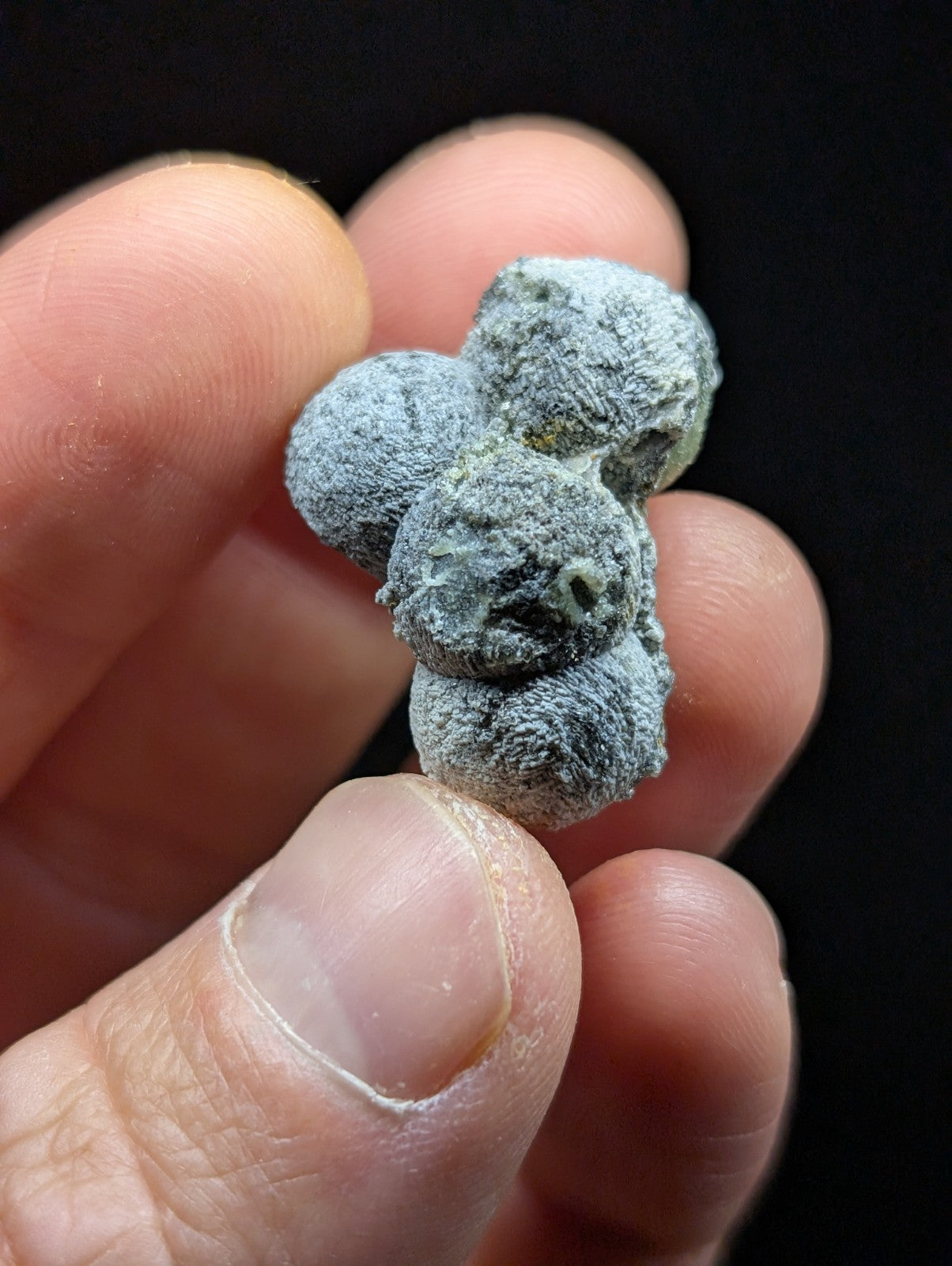 A gray, fuzzy, clustered mineral specimen held between fingers.