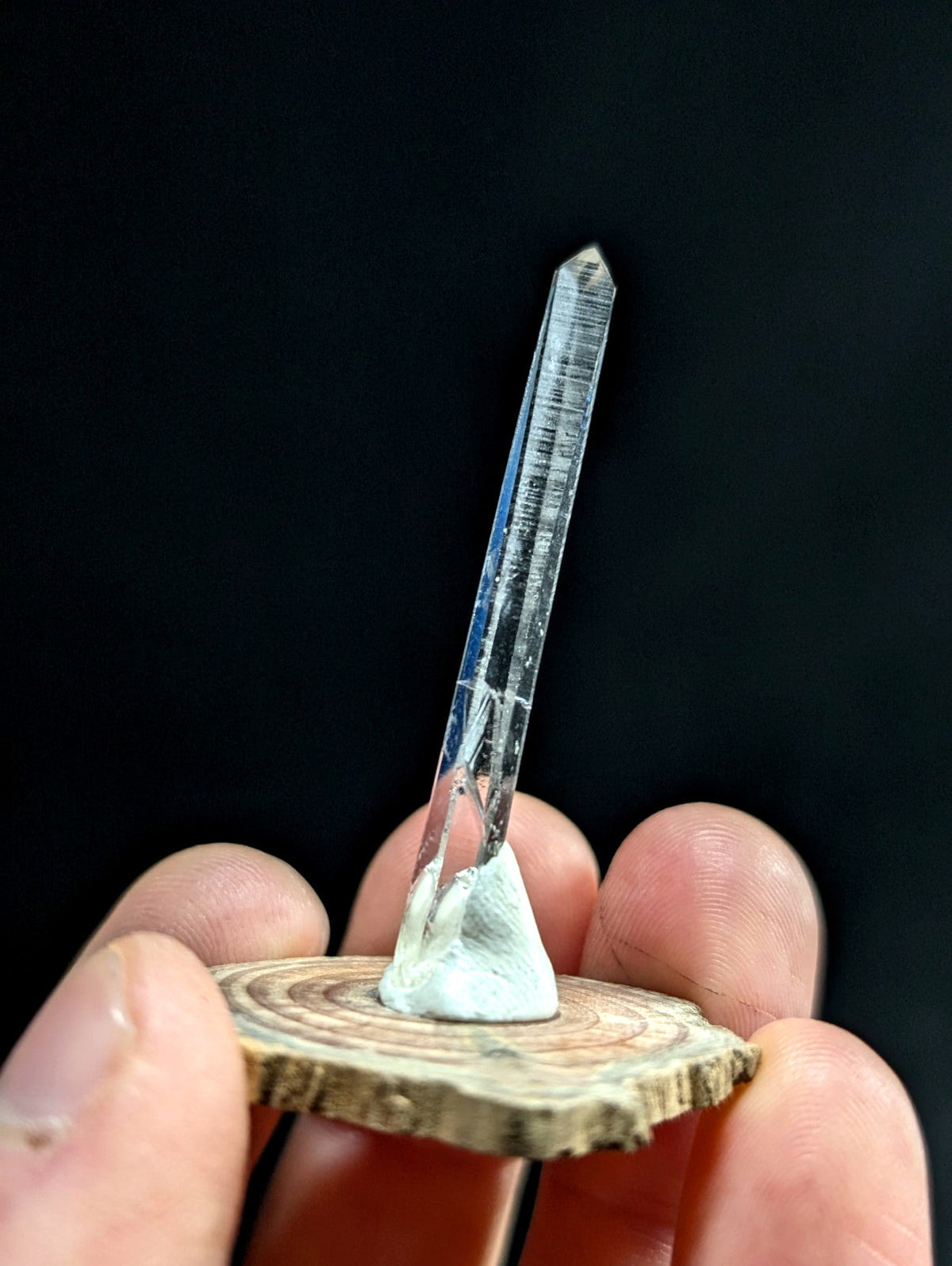A clear, faceted quartz crystal with natural internal striations stands upright on a circular wooden slice base.