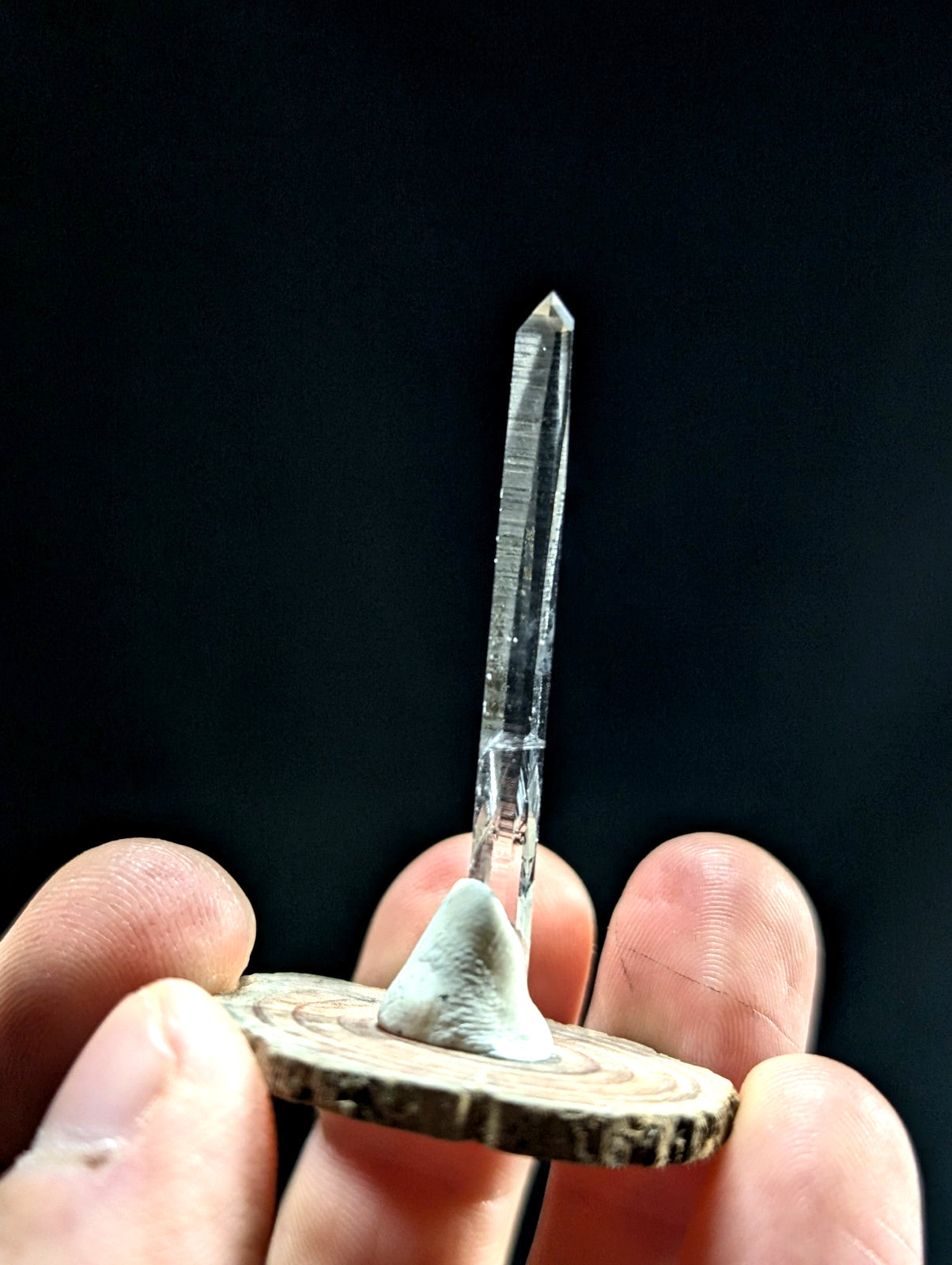A clear, transparent quartz crystal with a pointed tip and visible internal striations stands upright on a circular wooden base.