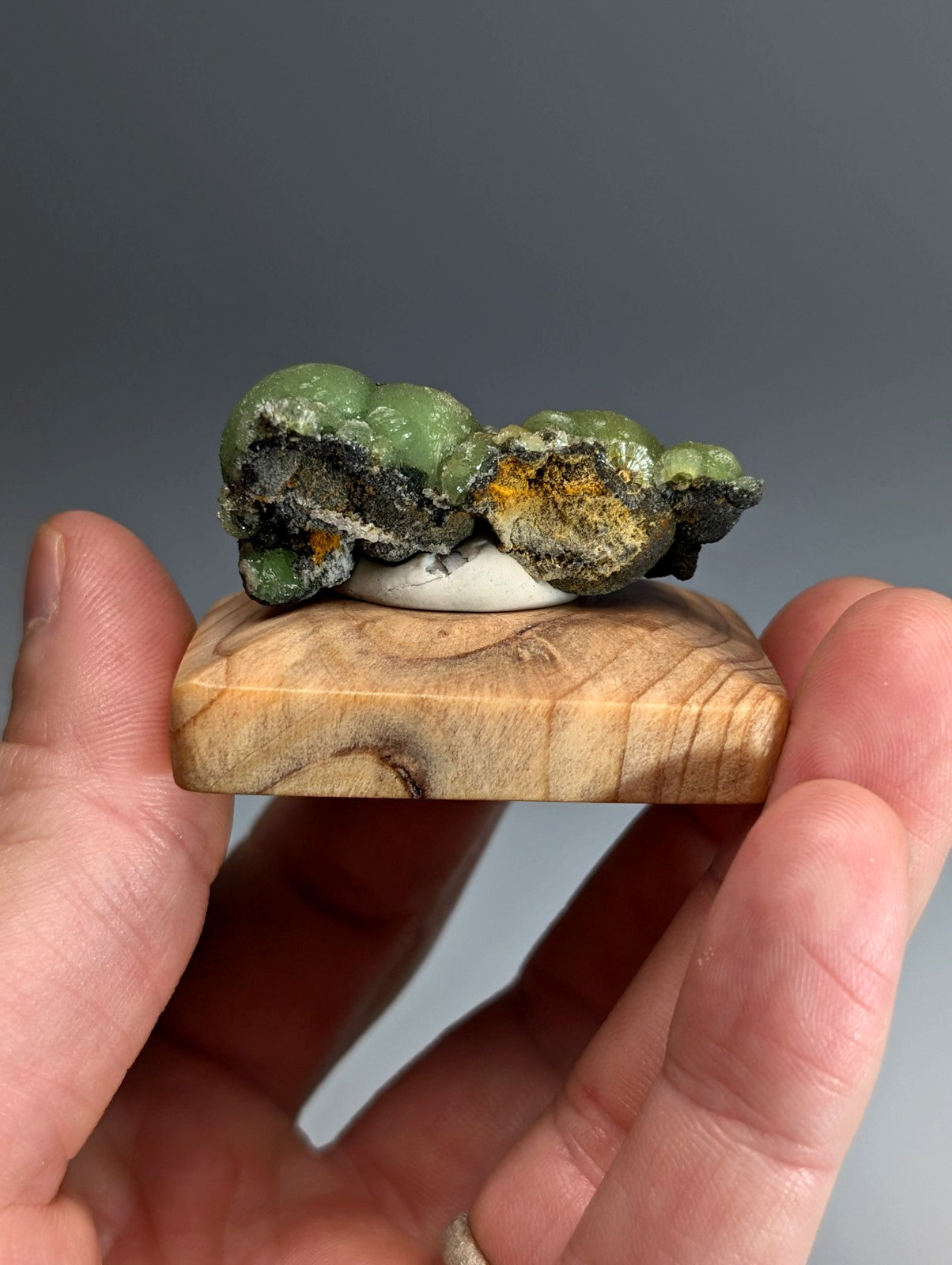 A hand holds a vibrant green and dark gray A+++ museum grade wavellite on a handmade wood mount from Arkansas
