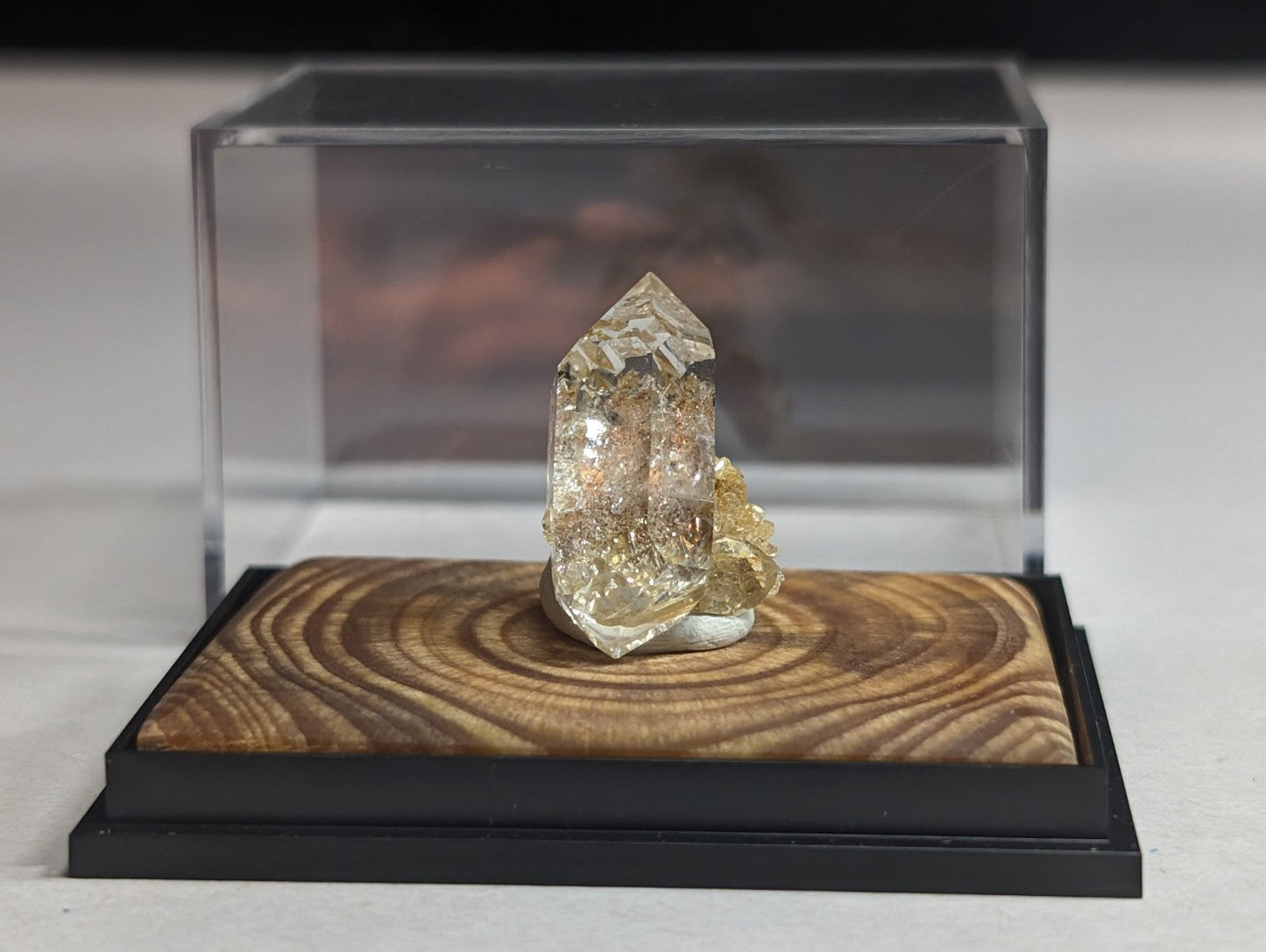 A clear, faceted quartz crystal with golden inclusions sits atop a wooden base within an acrylic display case.