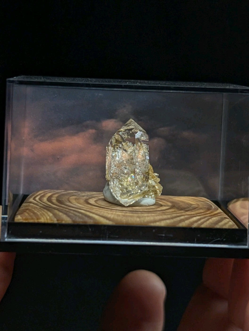 A clear, faceted quartz crystal with golden inclusions sits atop a wooden base inside a transparent display case.