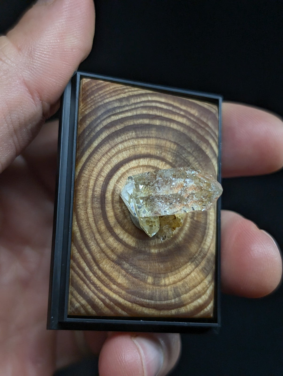 A clear, faceted quartz crystal with subtle golden inclusions rests atop a circular wood grain display within a sleek black frame.