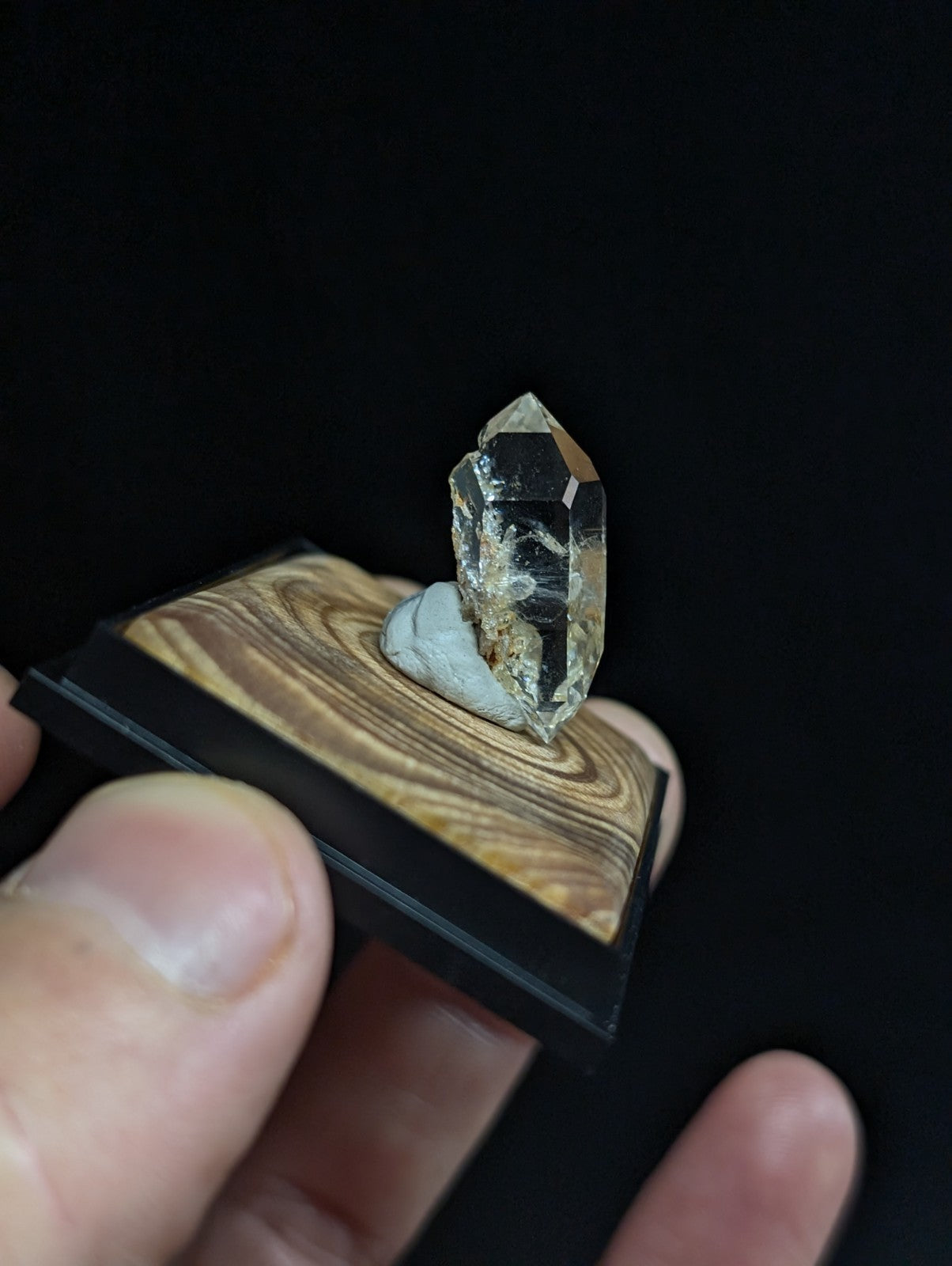 A clear, faceted quartz crystal with subtle internal inclusions stands upright on a polished wooden base framed by black material.