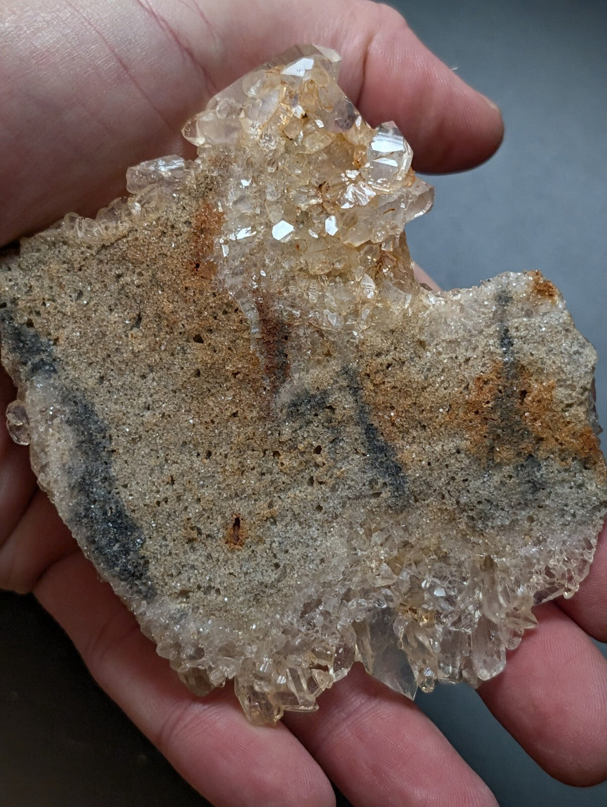 A hand holds a natural quartz crystal formation with translucent, faceted crystals growing from a rough, sandy matrix of earthy brown and gray tones.