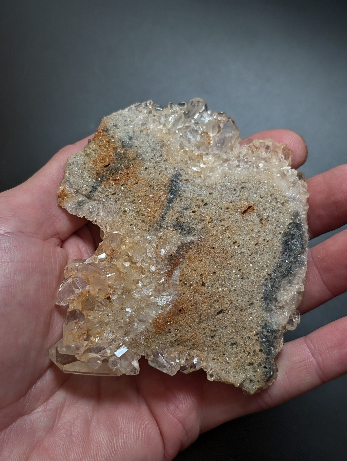 A hand holds a natural quartz geode with translucent, crystalline formations and earthy brown and gray mineral inclusions.