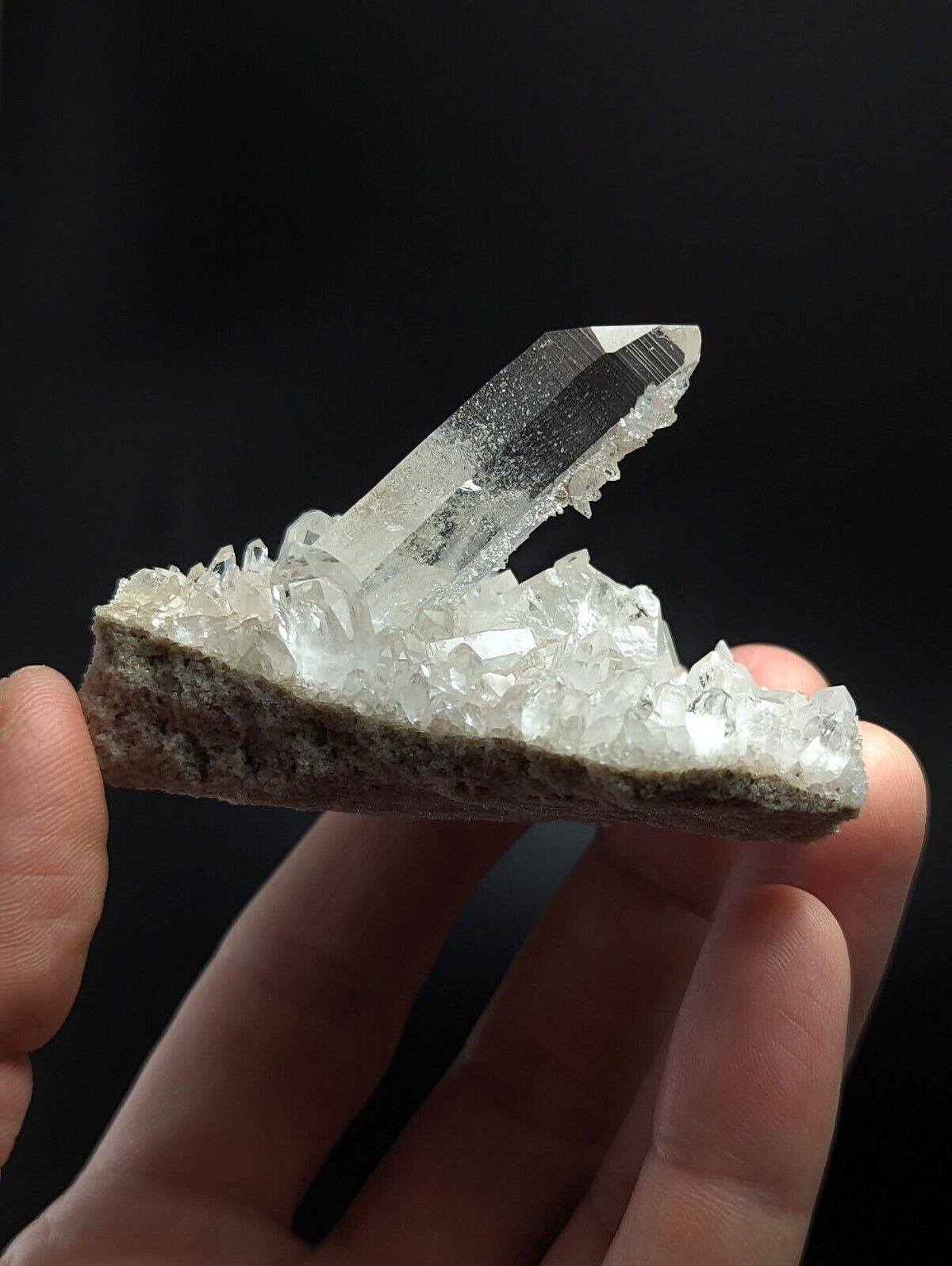 A clear quartz crystal cluster with a prominent, sharp-pointed central spike emerging from a bed of smaller translucent crystals embedded in a rough, earthy matrix.
