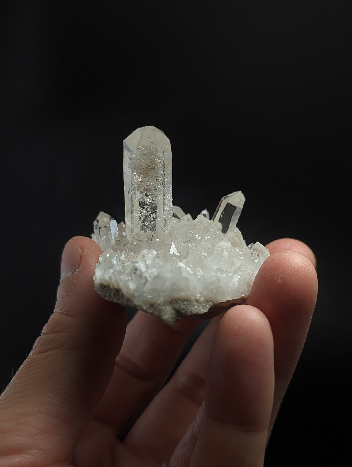 A clear quartz crystal cluster with a prominent central column and smaller surrounding crystals, held in hand against a dark background.