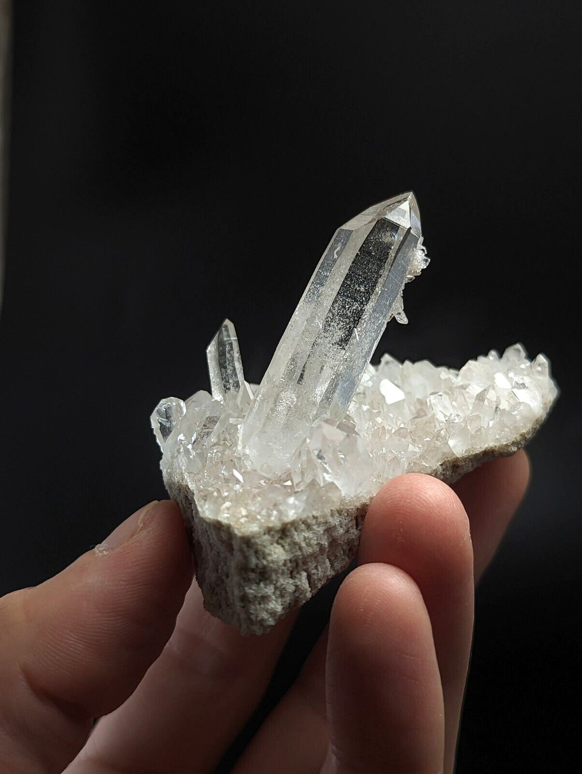 A clear quartz crystal cluster with a prominent central point and smaller crystals radiating outward, set in a rough gray matrix.