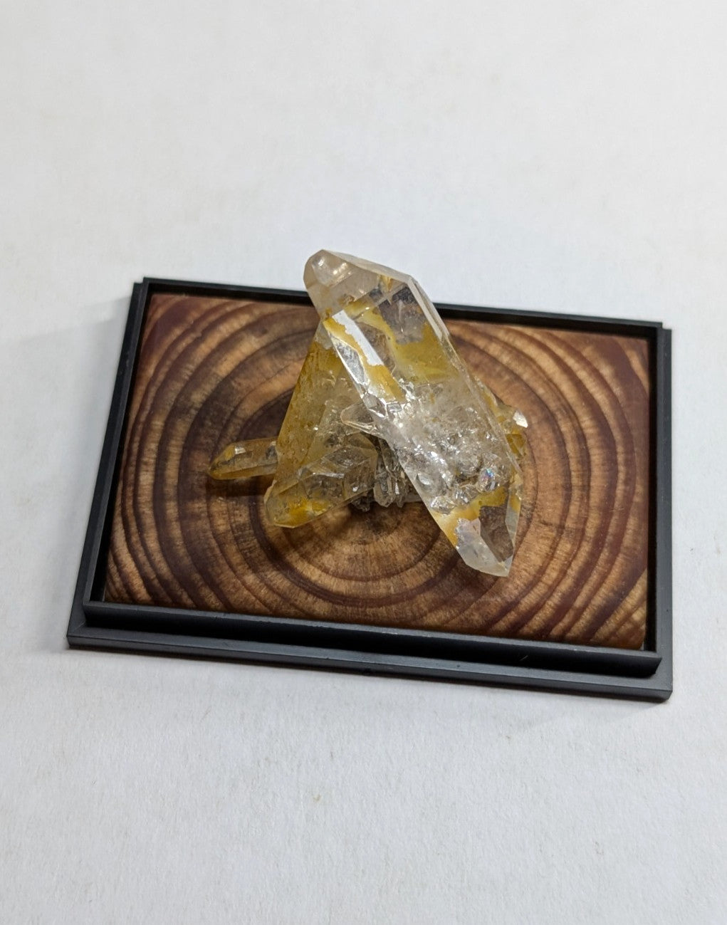 A translucent yellow and clear crystal cluster displayed in a black-framed wooden base with concentric ring patterns.