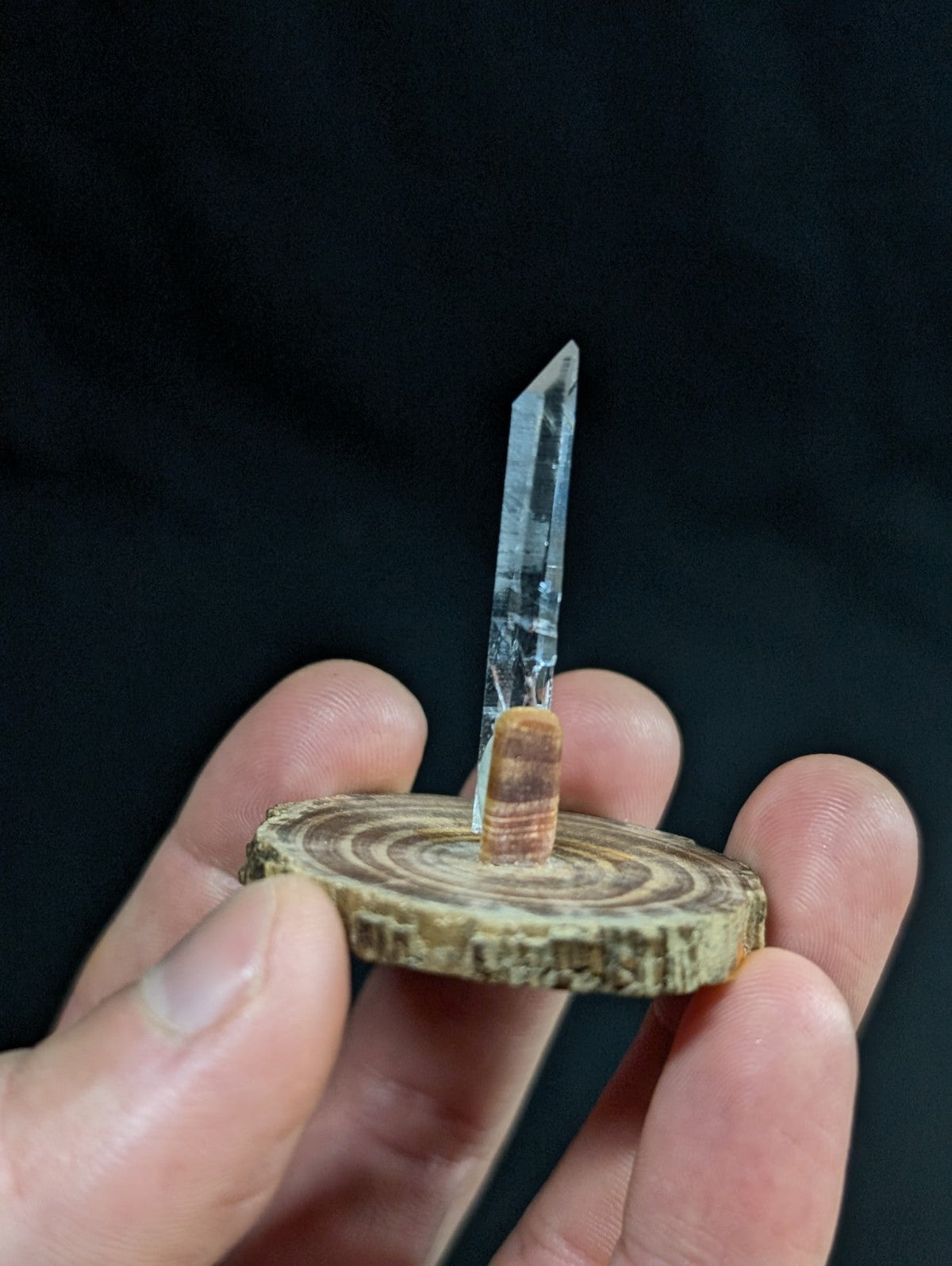 A clear quartz crystal stands upright on a rustic wooden slice, its pointed tip gleaming with natural translucency.