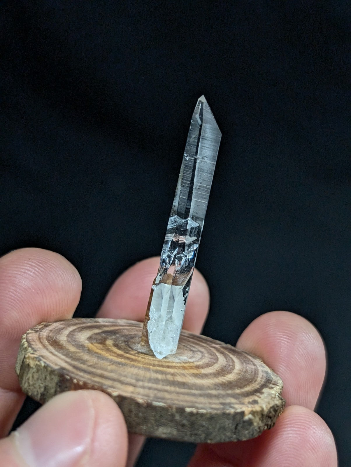 A clear quartz crystal with a pointed tip and visible internal striations, mounted on a rustic wooden slice.