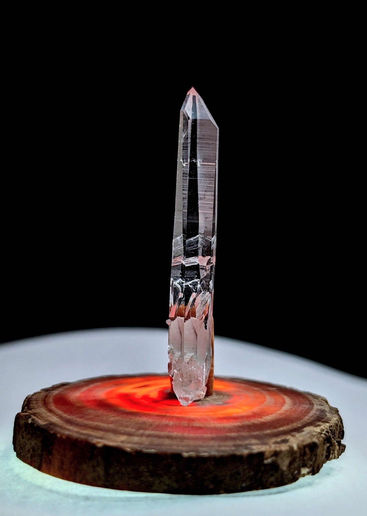 A tall, clear quartz crystal with a pointed tip and subtle internal striations stands upright on a circular wooden base.