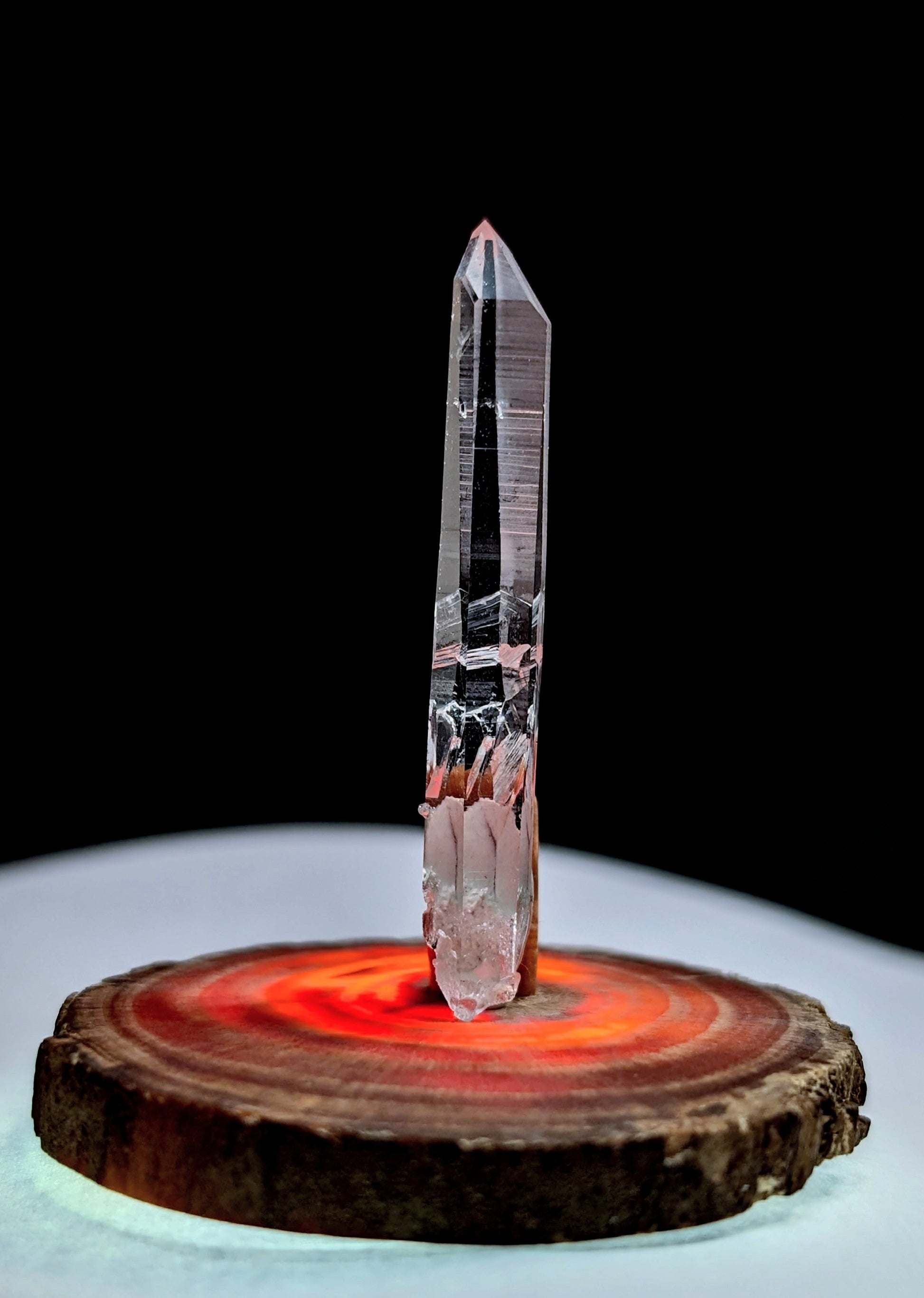 A tall, clear quartz crystal with a pointed tip and subtle internal striations stands upright on a circular wooden base.