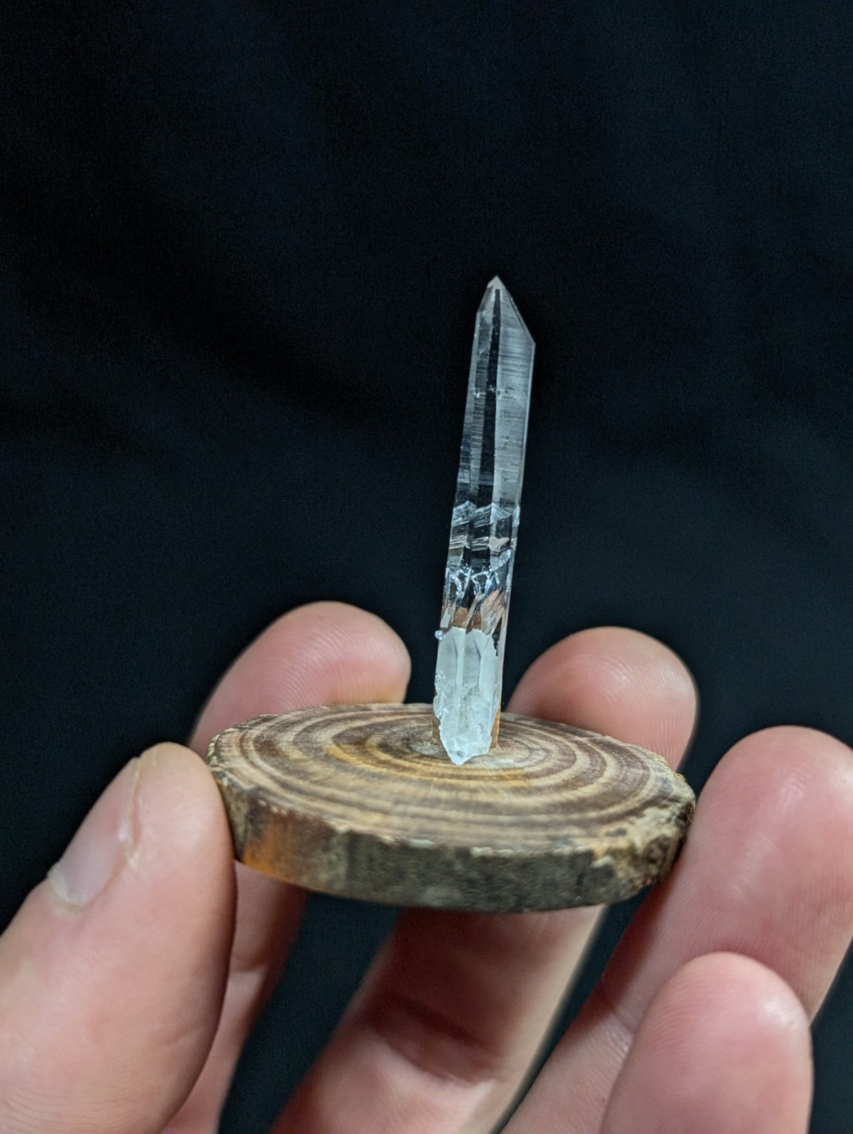 A clear, translucent quartz crystal with a pointed tip and subtle internal striations stands upright on a circular wooden base.
