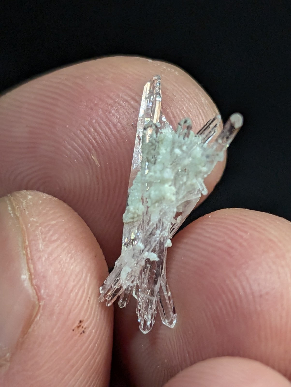 A delicate cluster of translucent, pale green crystals with sharp, glassy facets held gently between fingers.