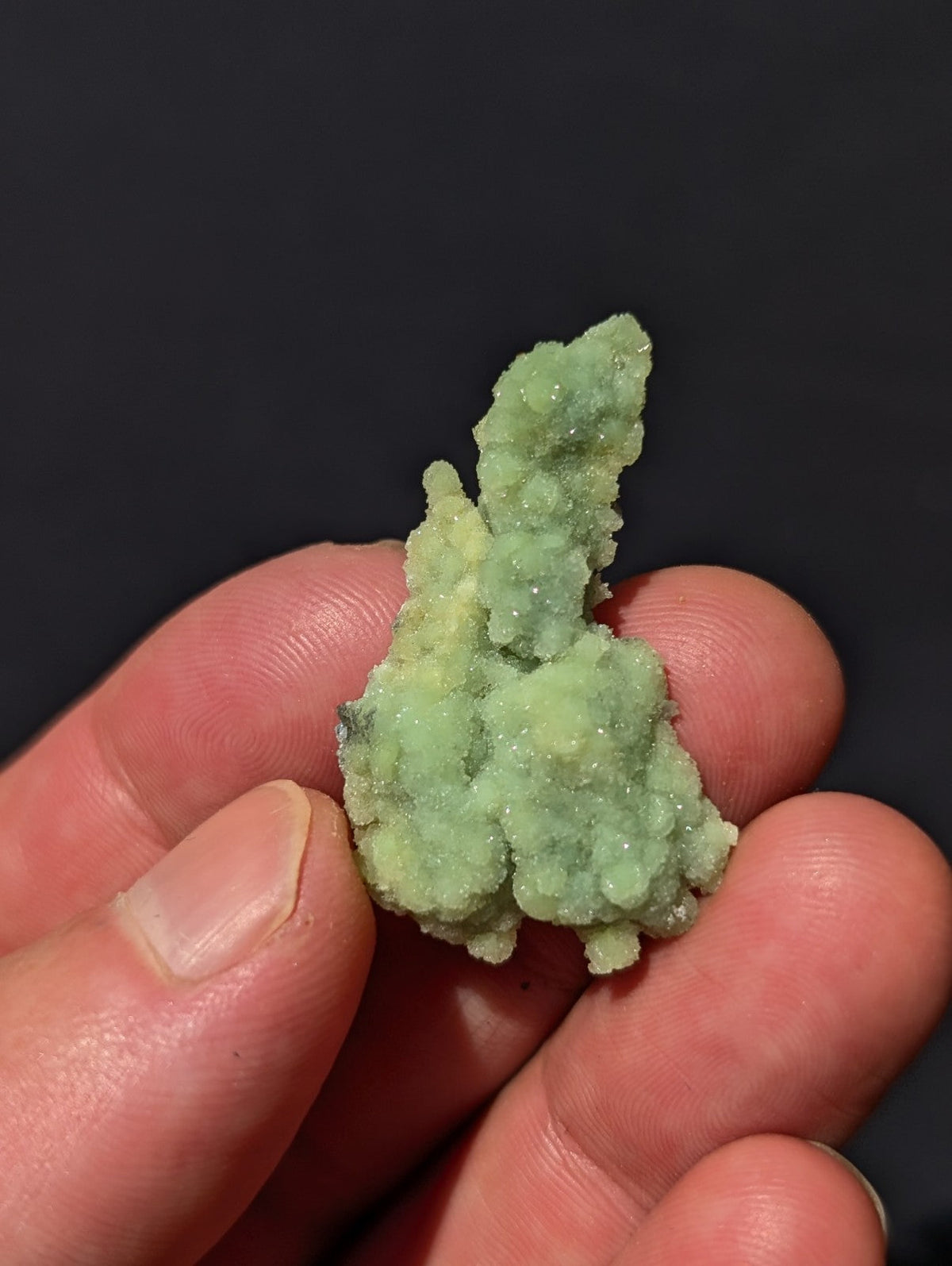 A delicate, pale green mineral specimen with a crystalline, textured surface held gently in someone’s hand.