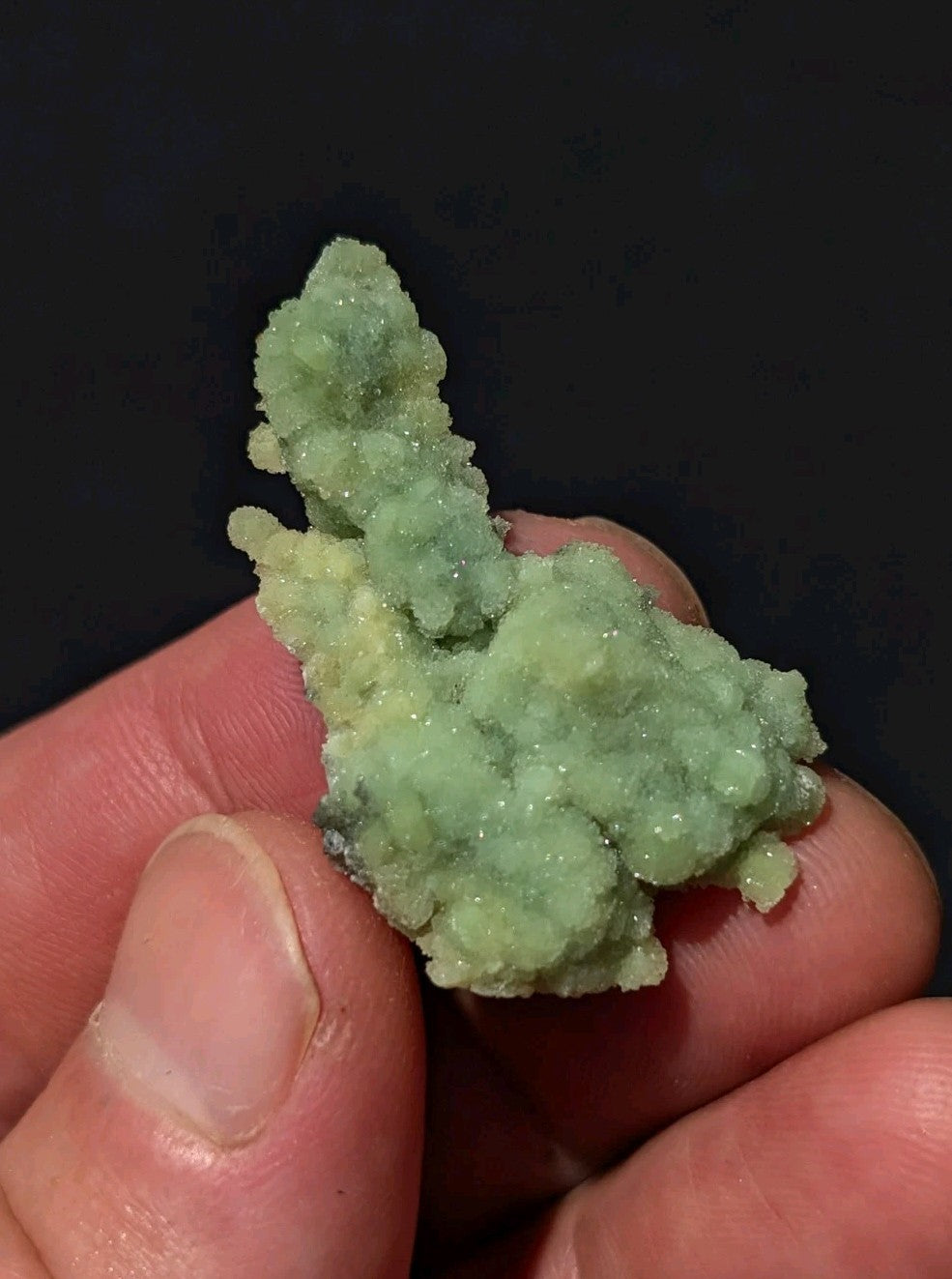 A pale green, crystalline mineral specimen with a rough, textured surface held gently in someone’s fingers.