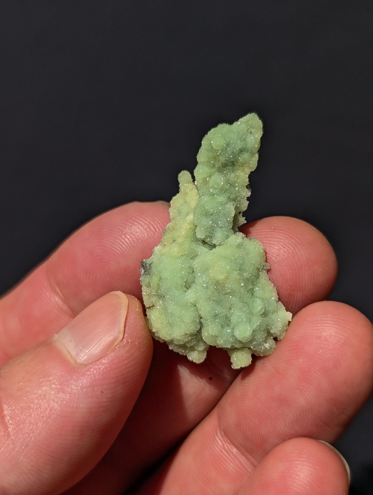 A pale green, crystalline mineral specimen with a rough, textured surface held gently in someone’s fingers.