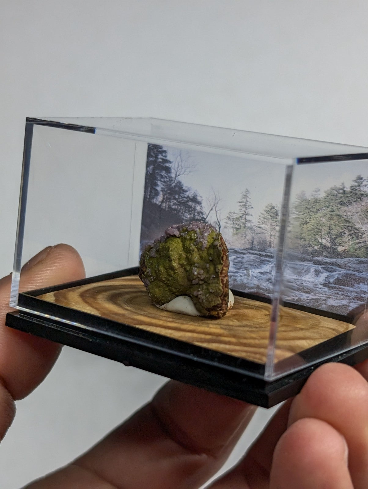 A mossy, earth-toned mineral specimen with a rough texture and subtle purple undertones is displayed in a clear acrylic display case.