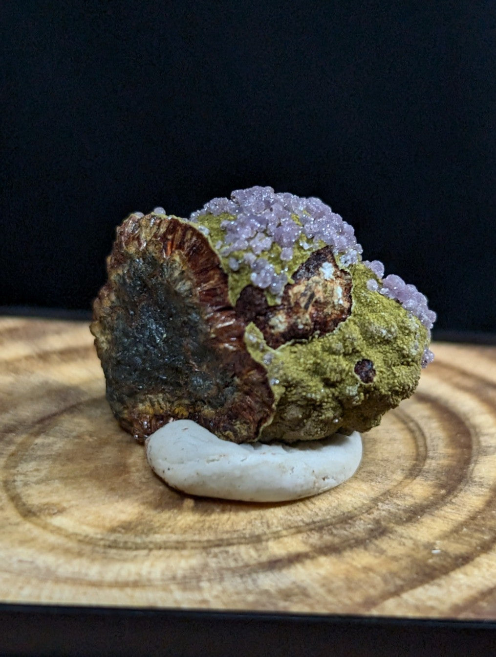 A striking mineral specimen featuring a rugged, earthy base with patches of green and brown, topped by clusters of delicate purple crystals.