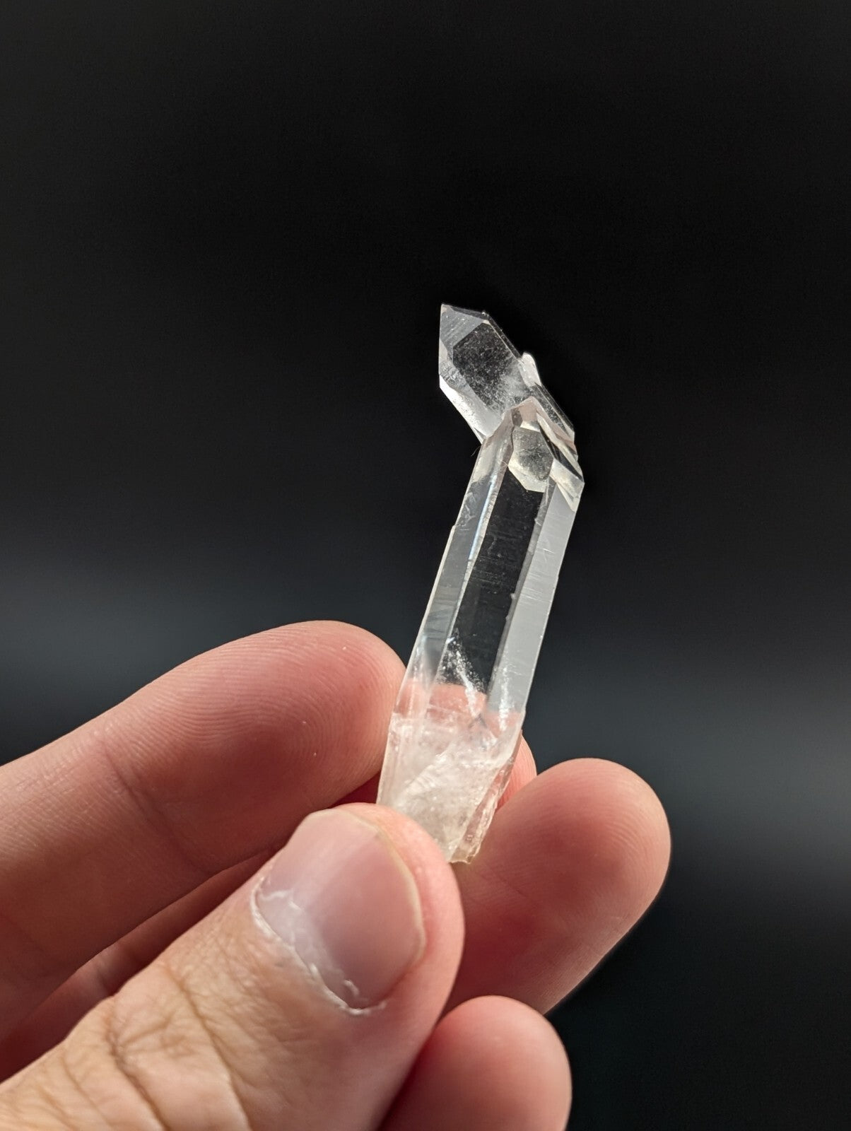 A clear quartz crystal with a pointed tip and faceted sides, held gently between fingers.
