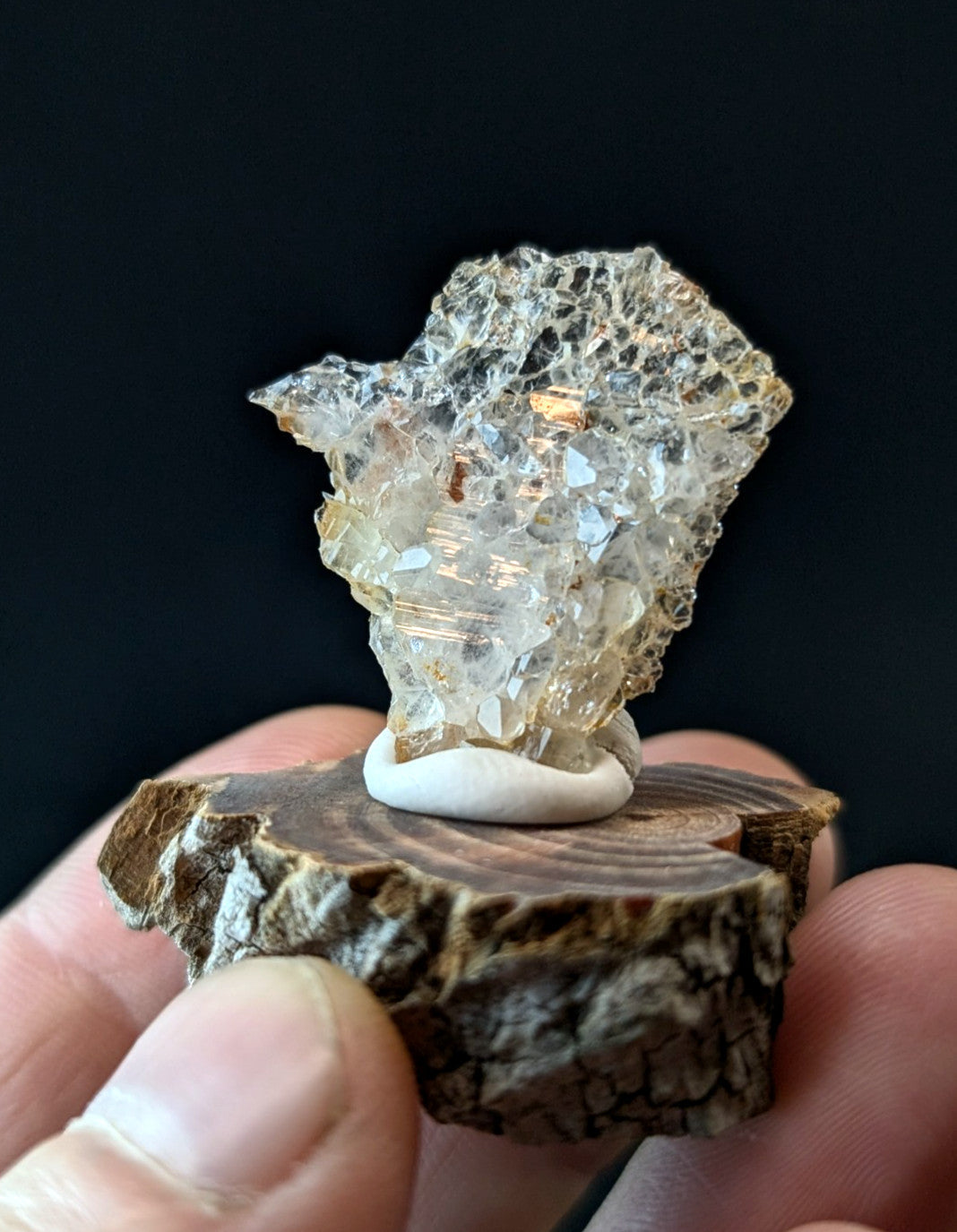 A translucent, crystalline mineral specimen with a rough, faceted surface and golden-yellow inclusions, displayed on a small white ceramic pedestal atop a rustic wooden slice.