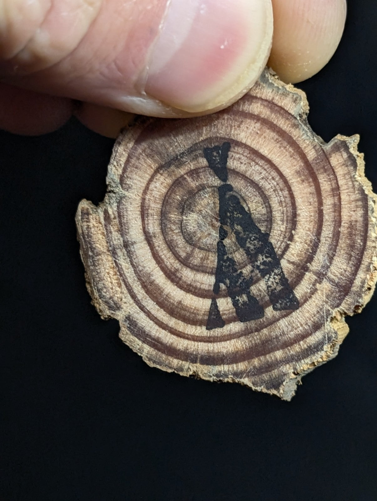 A rustic, circular wooden slice with natural concentric rings and a black inked silhouette of an arrowhead design.