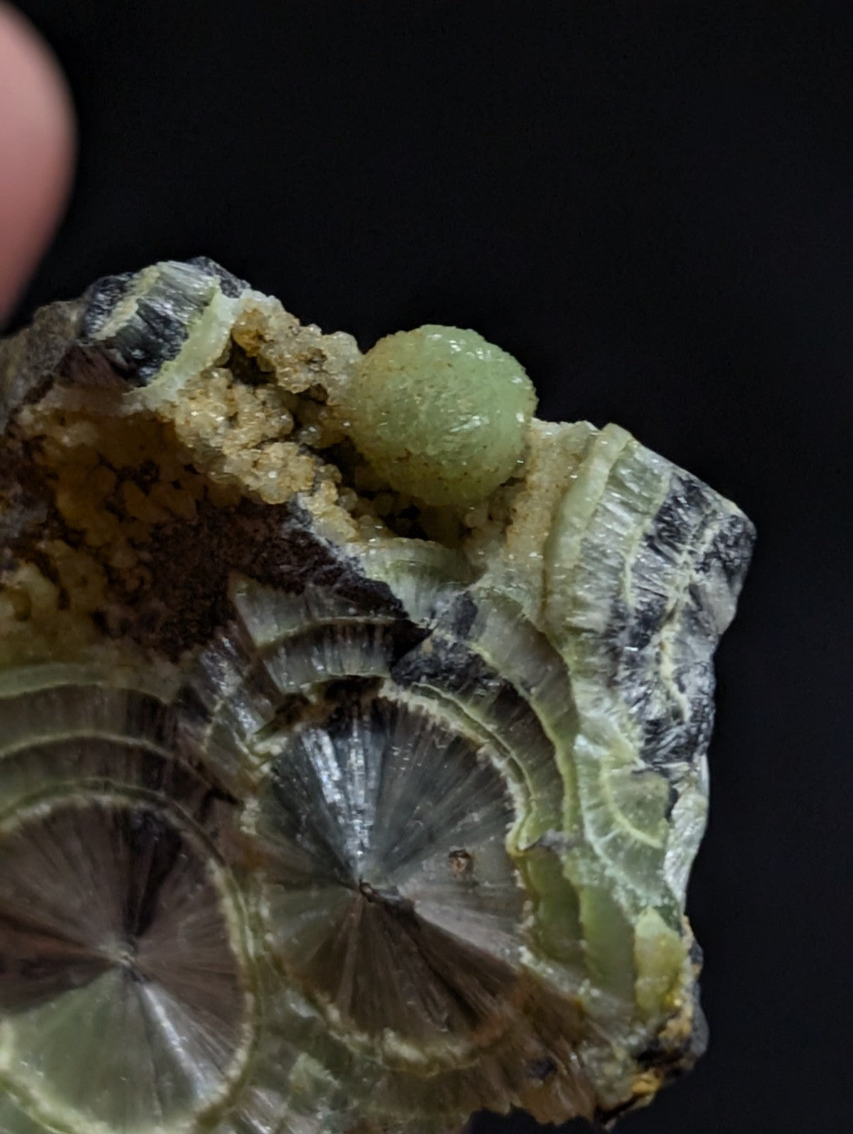 A green, spherical mineral specimen nestled atop a layered, translucent rock with concentric circular patterns.