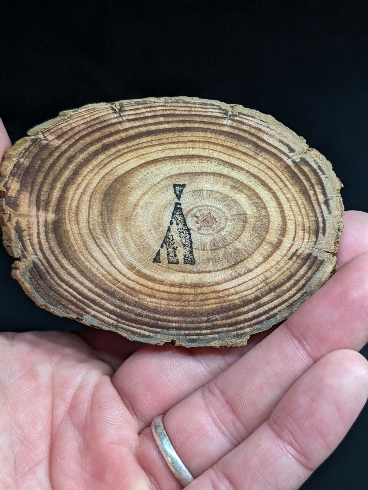 A rustic, oval-shaped wooden slice with natural concentric rings and a dark charcoal-burnt silhouette of a teepee at its center.