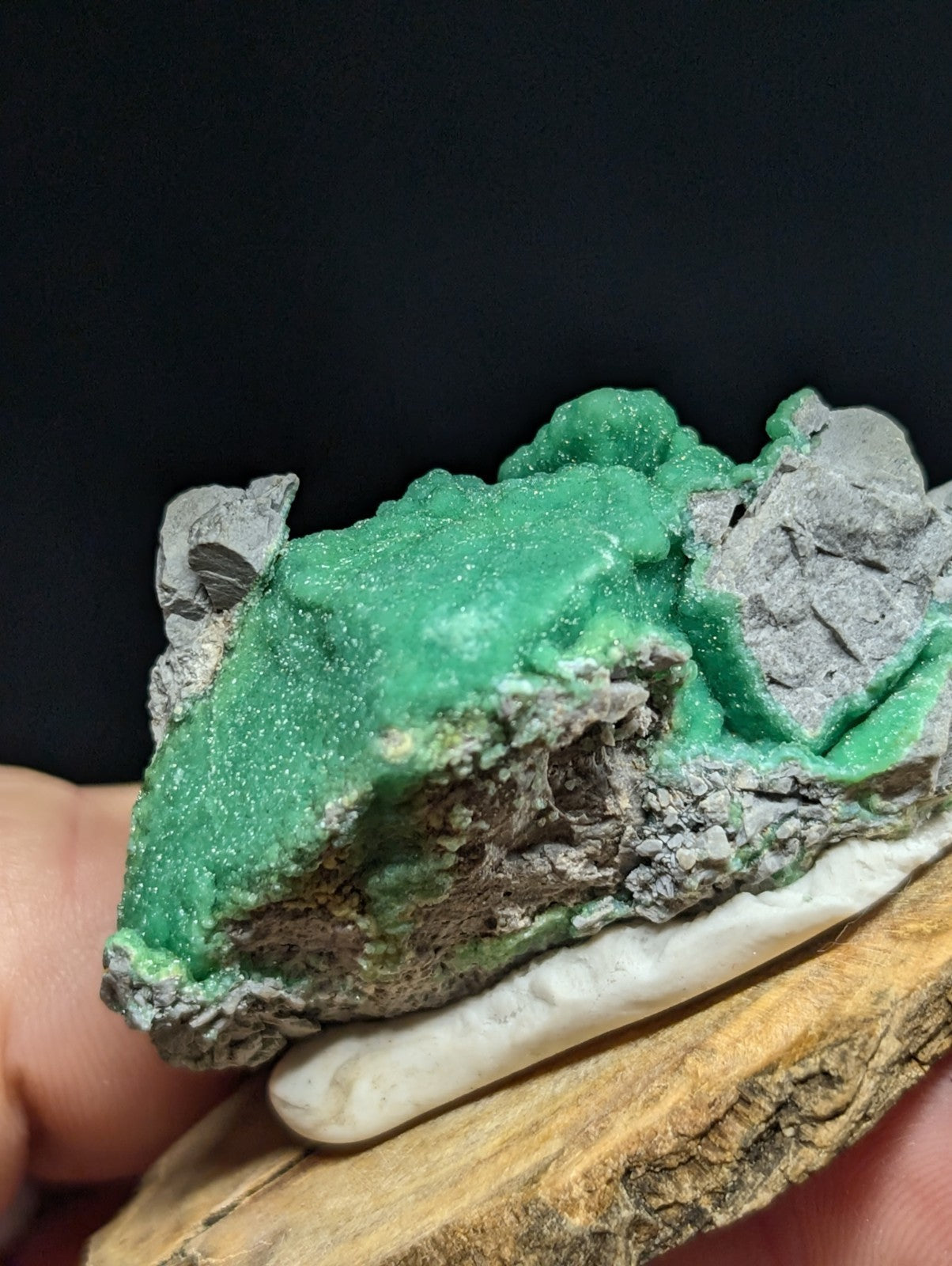 A vibrant green, glittering mineral specimen with a rough, textured surface set against gray rock matrix.