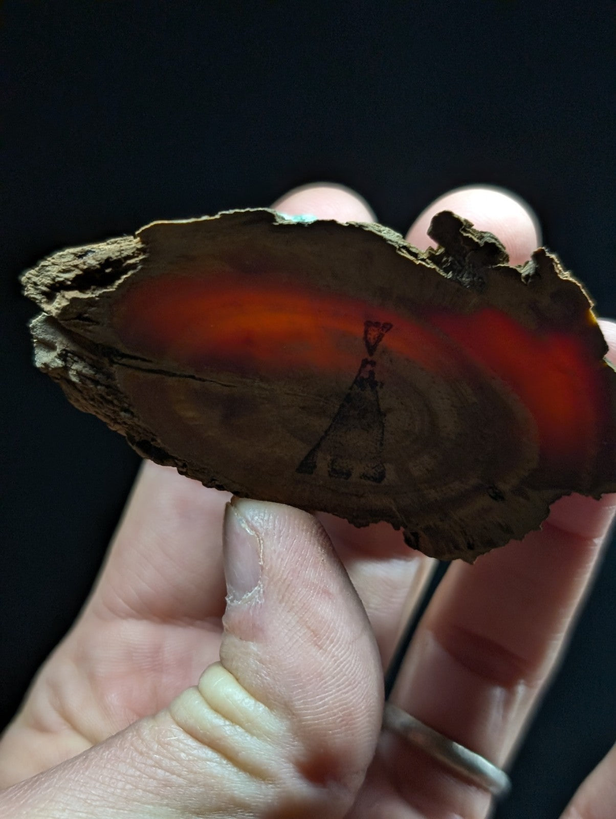 A hand holds a rustic, irregularly shaped piece of amber with vibrant orange and reddish hues radiating from its center, featuring an engraved silhouette of a teepee.